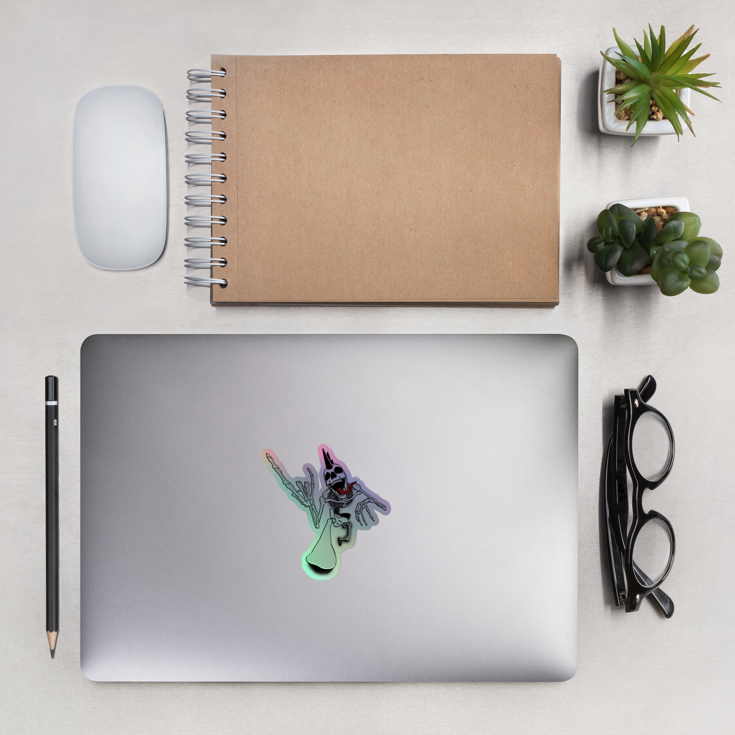 Laptop with a a holographic sticker depicting a punk rock skeleton with a colorful, shimmering, holographic outline, holding up heavy metal horns with a notebook, mouse, pencil, glasses, and small plants on a light surface.