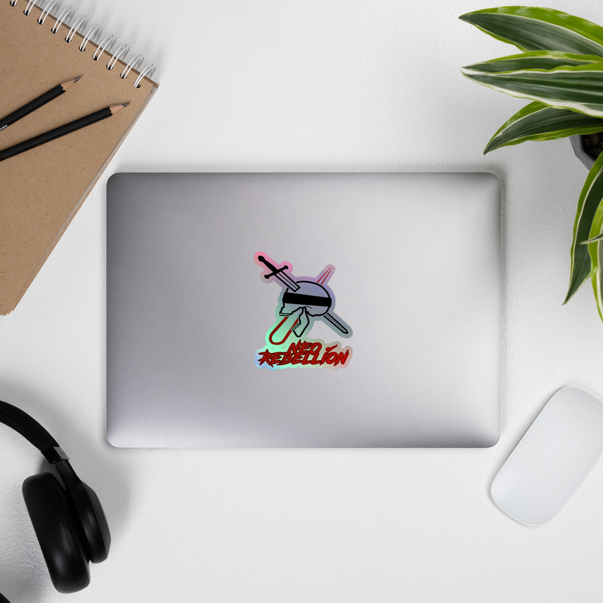 Laptop with a sticker with a skull, sword, laser and 'Neo Rebellion' text on a white background on a white surface with a plant and office supplies.