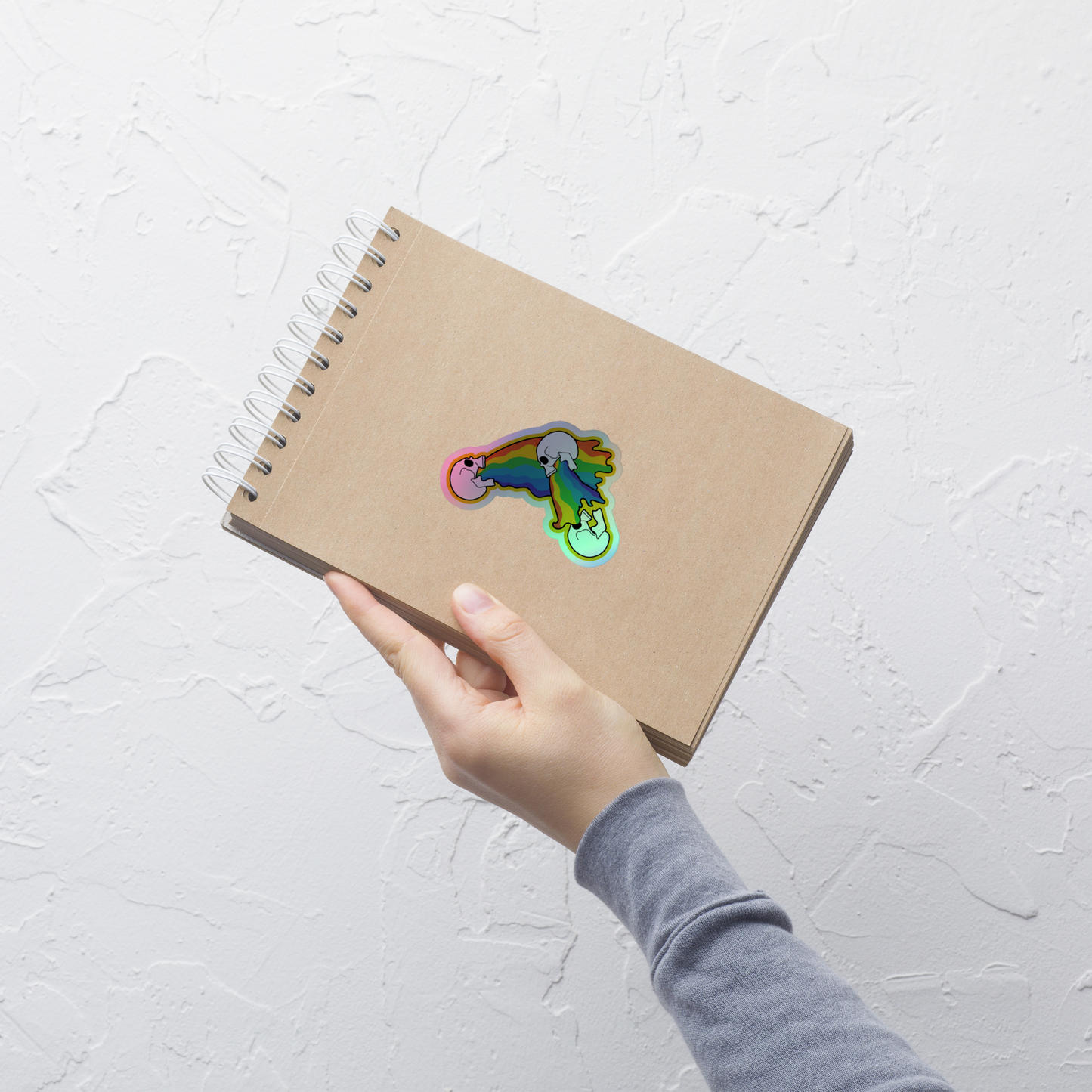 Hand holding a brown notebook with a colorful sticker of skulls vomiting a rainbow on a white background on a white surface design on a white textured background