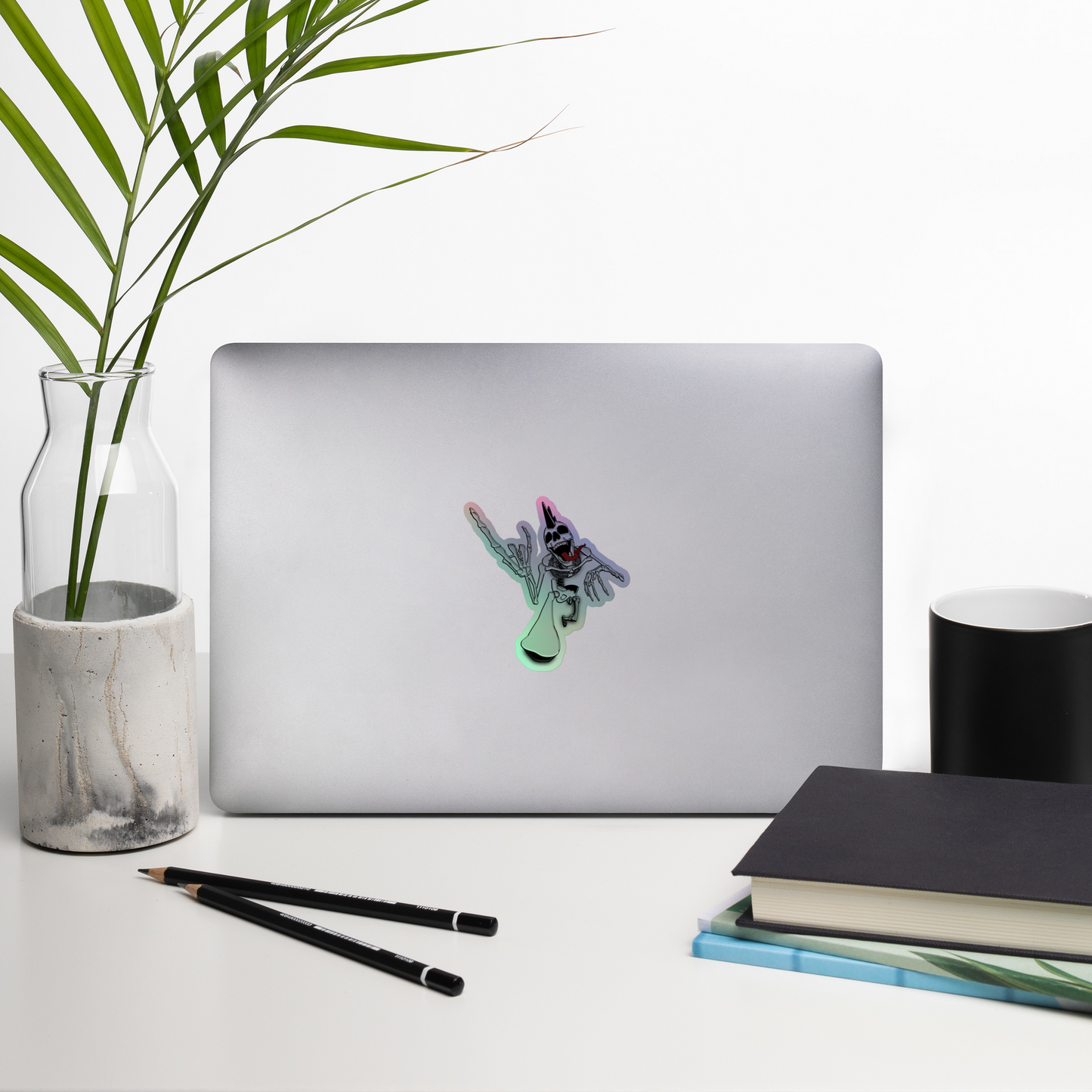 Laptop with a decorative holographic sticker of a punk rock skeleton holding up heavy metal horns on a desk with a plant, pencils, and books.