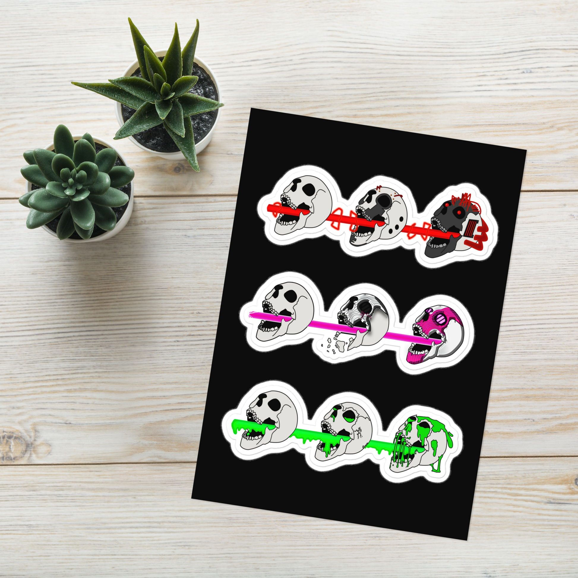 Set of three sticker designs featuring cyberpunk and horror themed skulls shooting different colored lasers on a black background, placed on a wooden surface with plants.