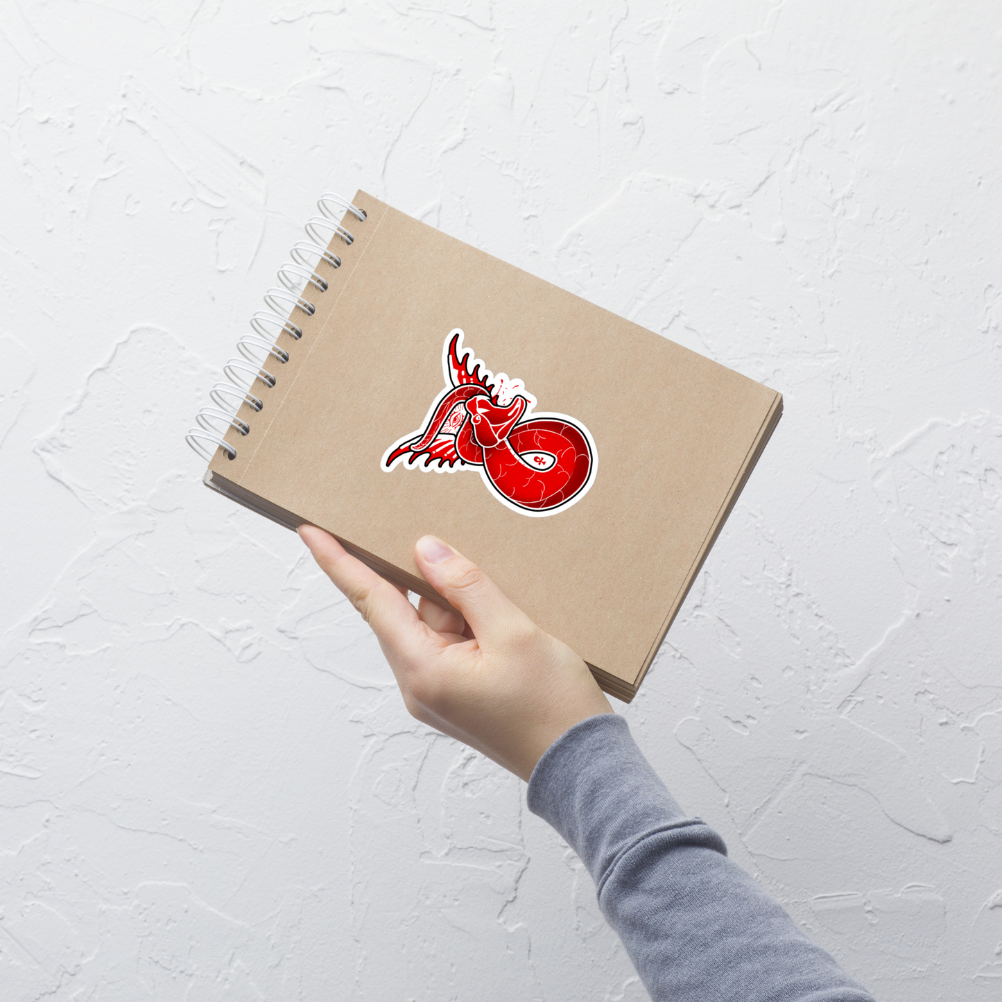 Hand holding a brown notebook with a red and white sticker on a light gray background