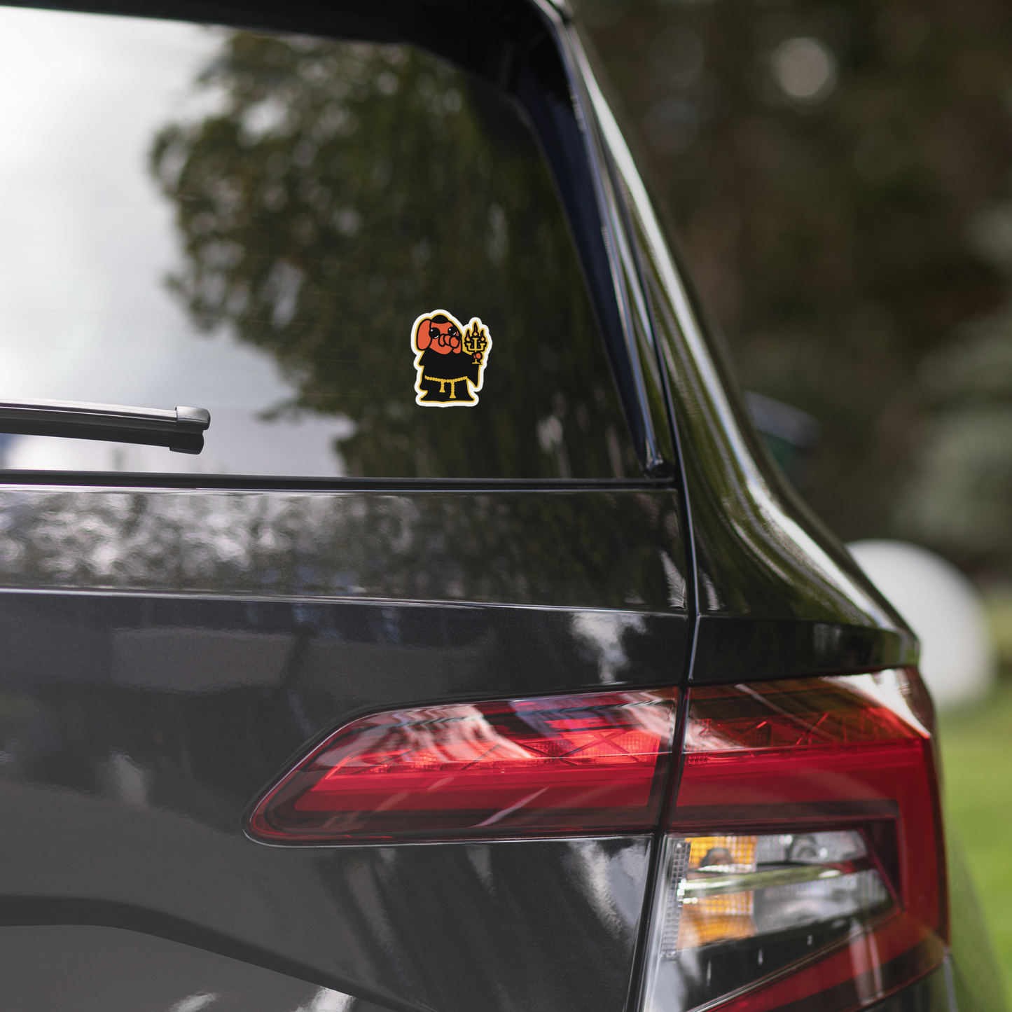 Car with an orange cartoon dog, black robe, and holding a candelabra on a white background sticker on the rear window, blurred greenery in the background