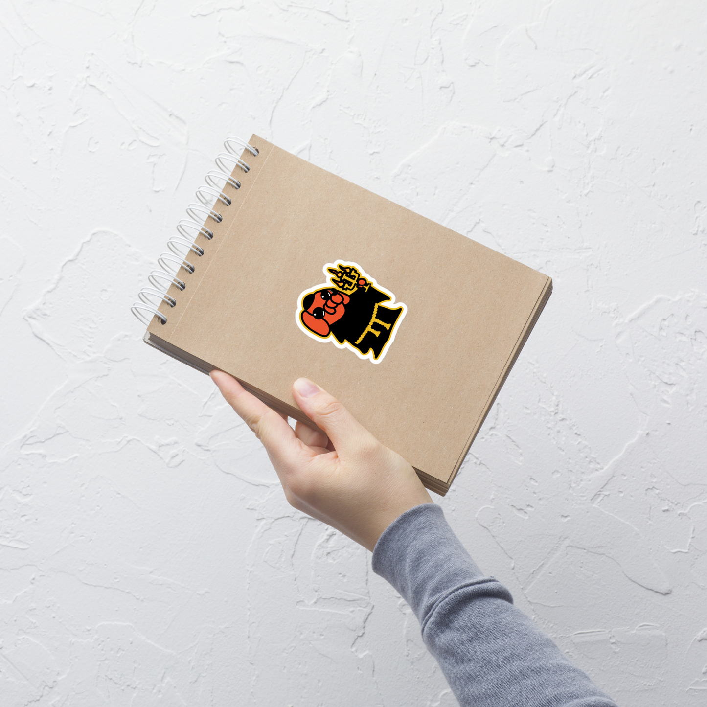 Hand holding a brown notebook with an orange cartoon dog, black robe, and holding a candelabra sticker on a white background in front of a light gray wall