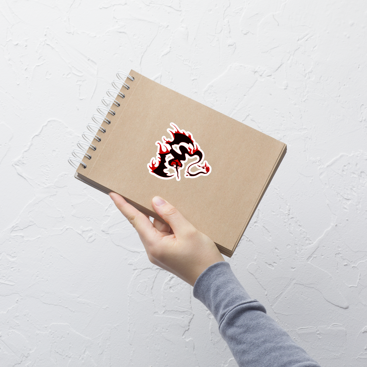 Hand holding a brown notebook with a black snake dragon with red flames on a white background sticker in front of a white wall