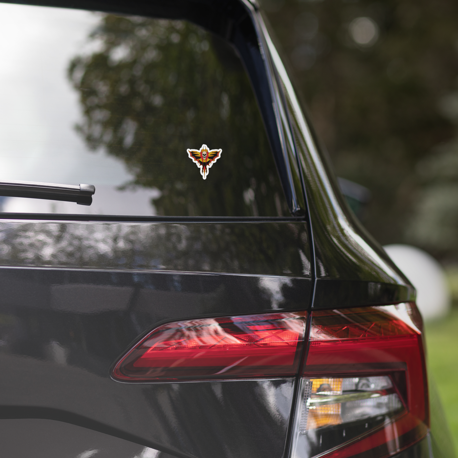 Car with a small sticker with a tattoo skull moth design featuring wings on a white background on the rear window, surrounded by trees.