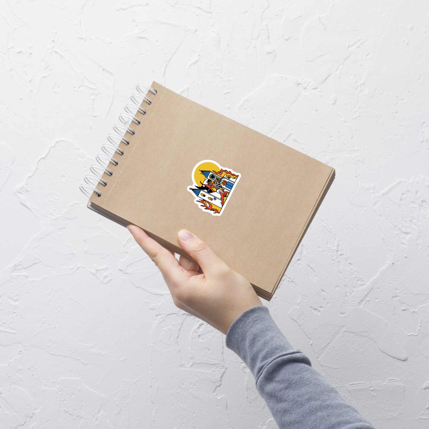 Hand holding a brown notebook with a colorful sticker with a colorful illustration of a giant monster attacking a flaming church full of teddy bears sticker on a light gray background