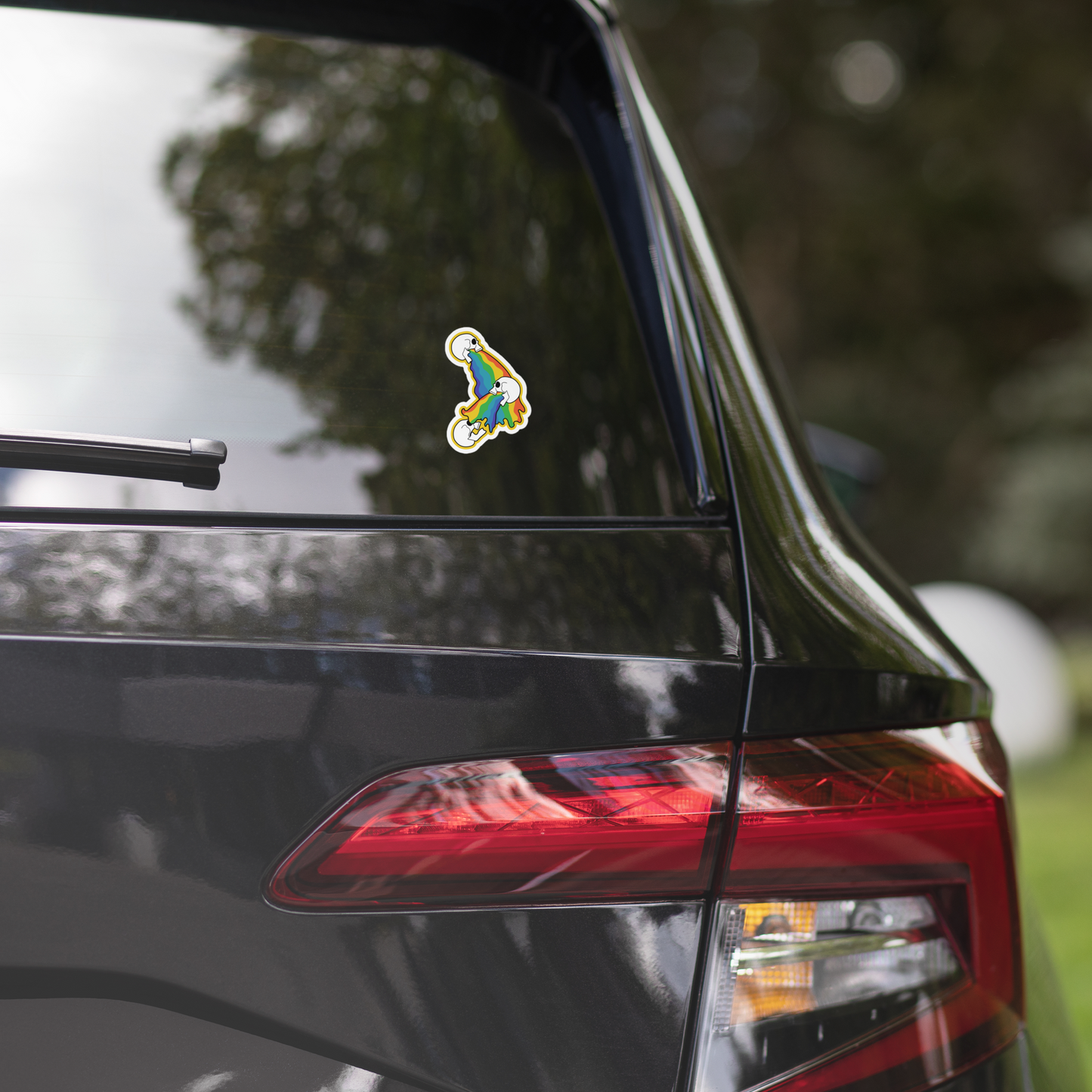 Colorful rainbow sticker of 3 skulls vomiting on the rear window of a black car with blurred greenery in the background