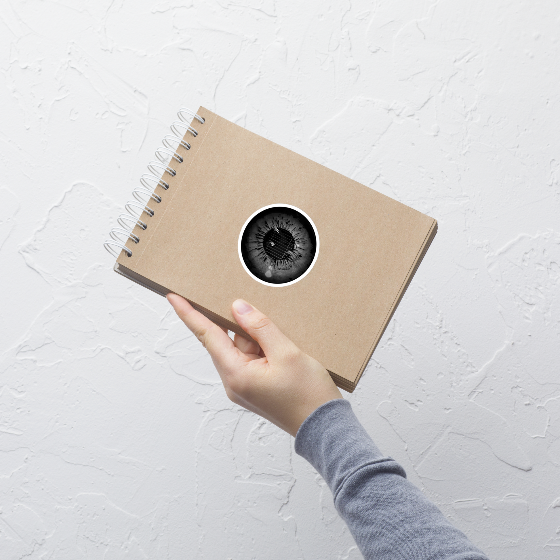 Hand holding a brown notebook with a circular black and white eye sticker with the grim reaper in a cage in a black metal style on a light gray textured background