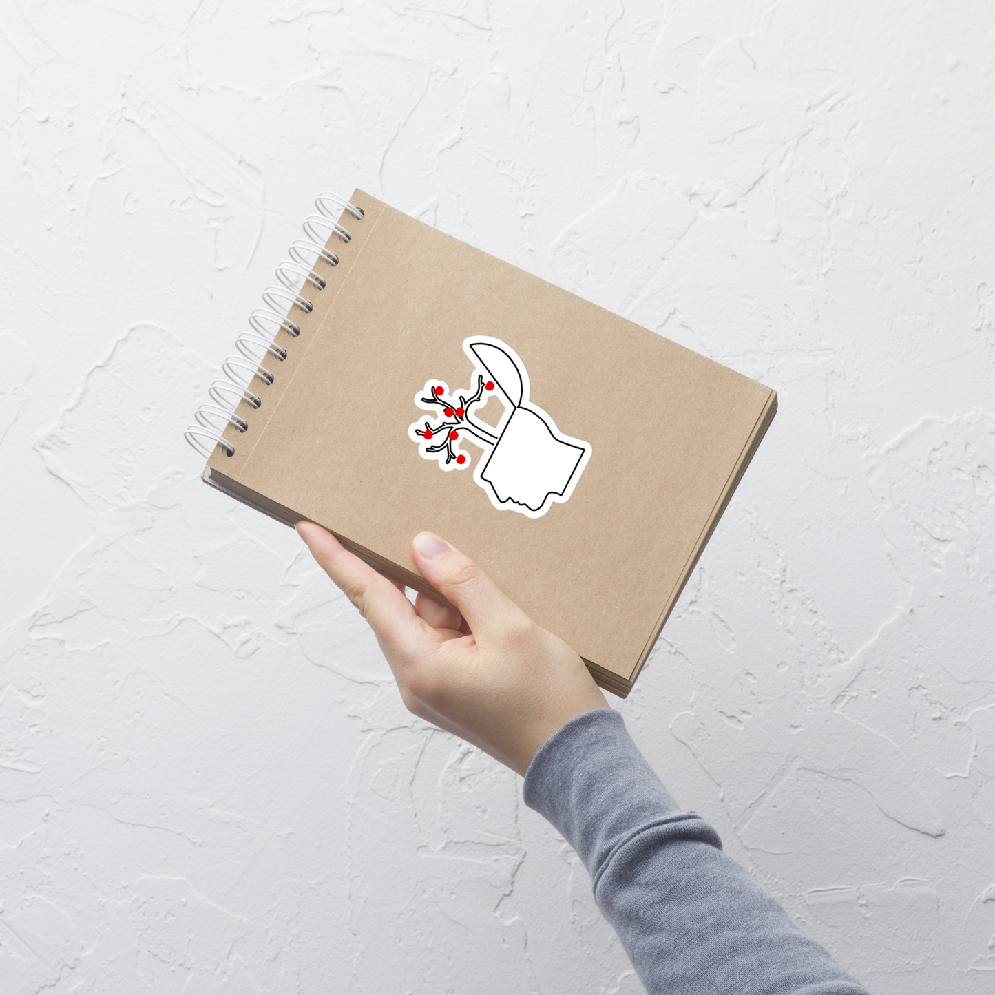 Hand holding a brown notebook with a sticker of an outline of a head with an open brain featuring branches with red apples on a white background on a light gray background