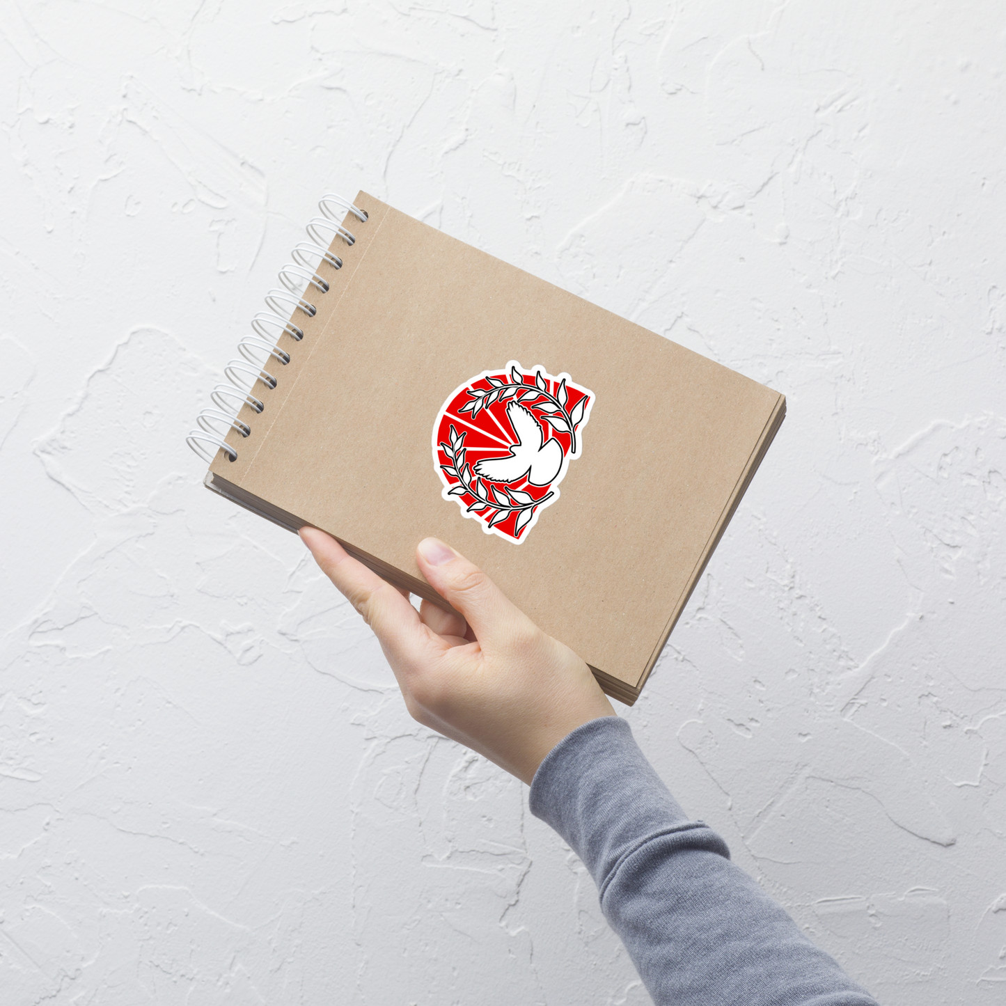 Hand holding a brown notebook with a red and white peace dove sticker emblem on a light gray background