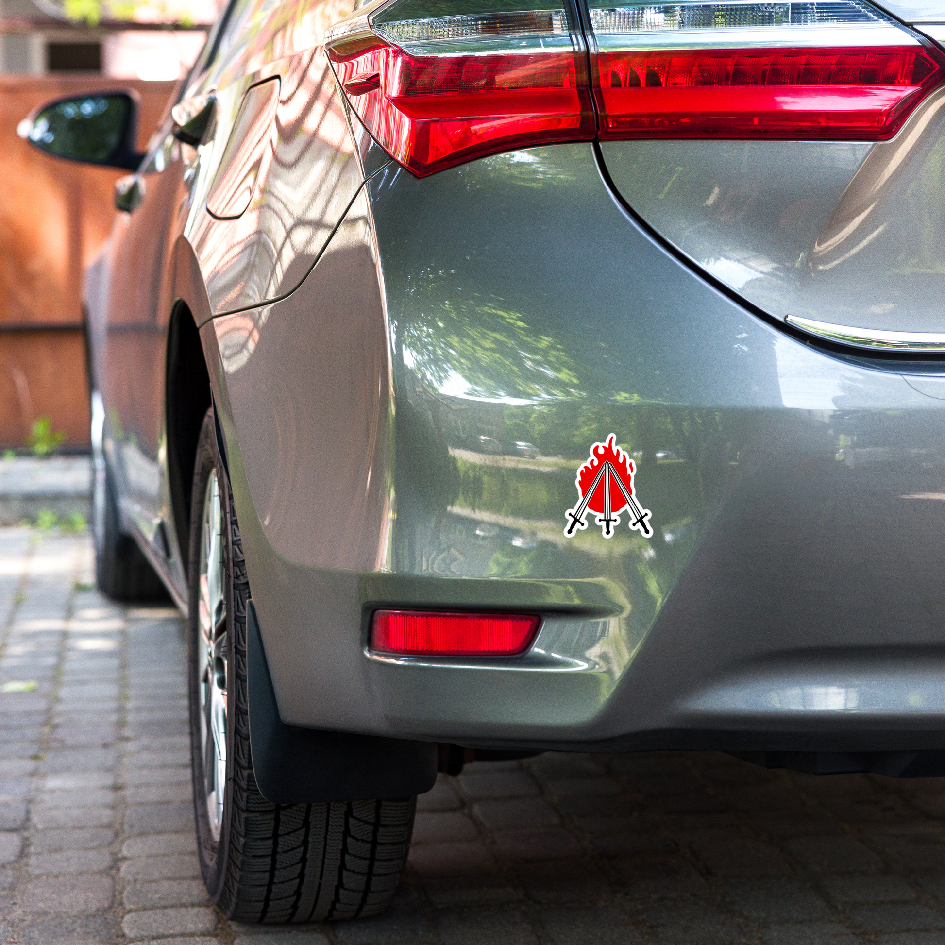 Sticker of three crossed swords in front of a red flame on a white background on the bottom bumper of a car on brick pavement
