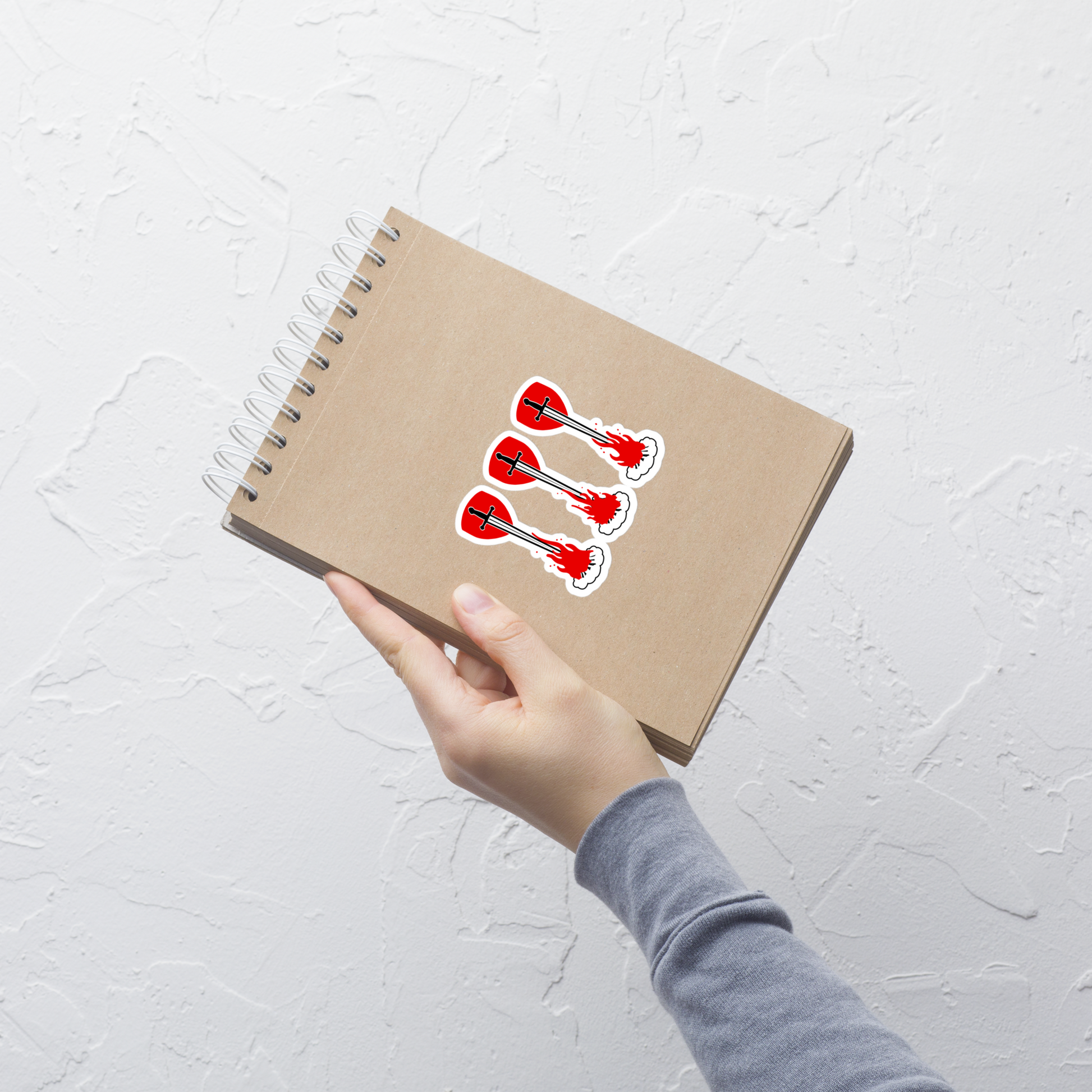 Hand holding a brown notebook with red, white, and black swords over campfire and shields sticker on a light gray background