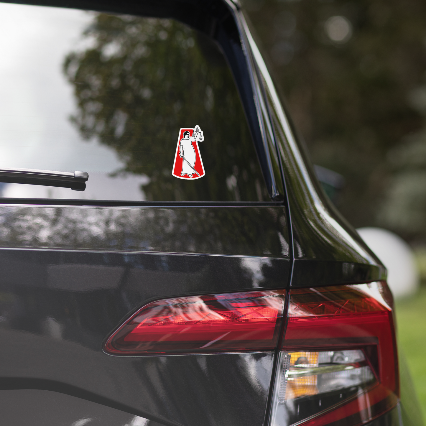 Car with a sticker of a lady justice figure holding a sword and scale on a red background on the back window.