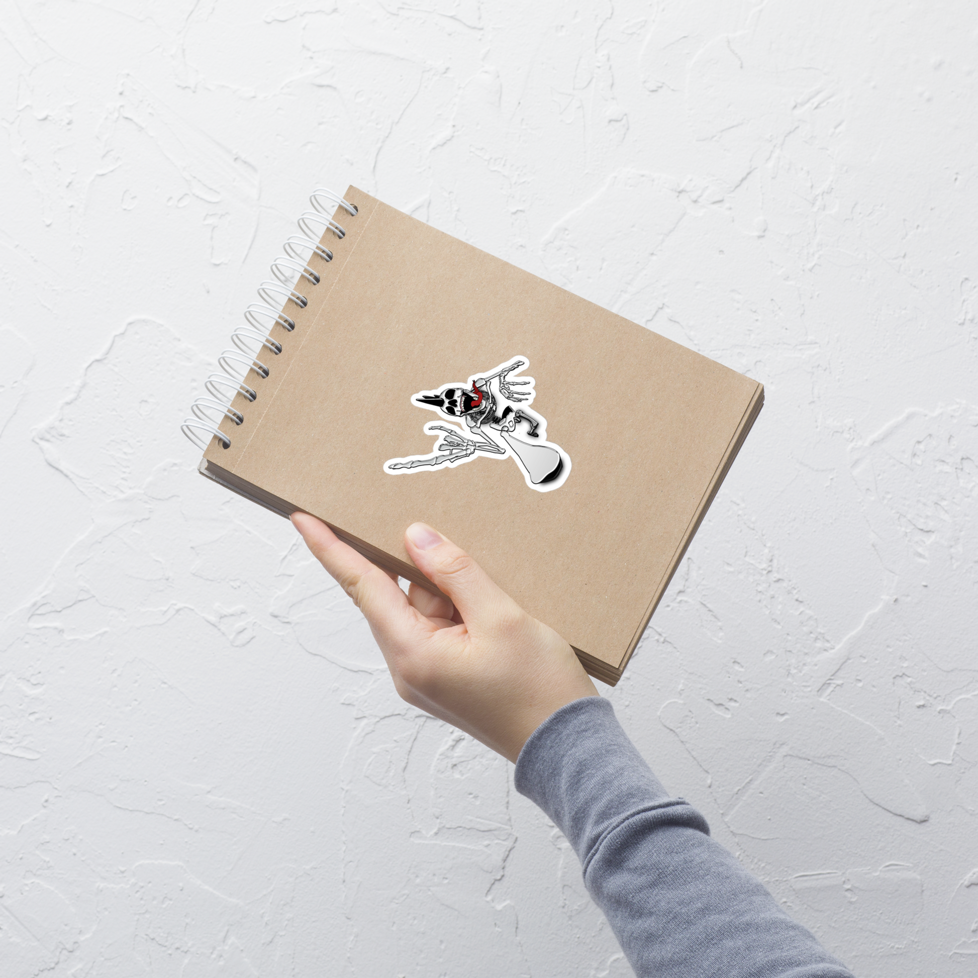Hand holding a brown notebook with a sticker of a heavy metal chrome skeletal character with a red tongue and a mohawk, on a white background on a light gray textured background