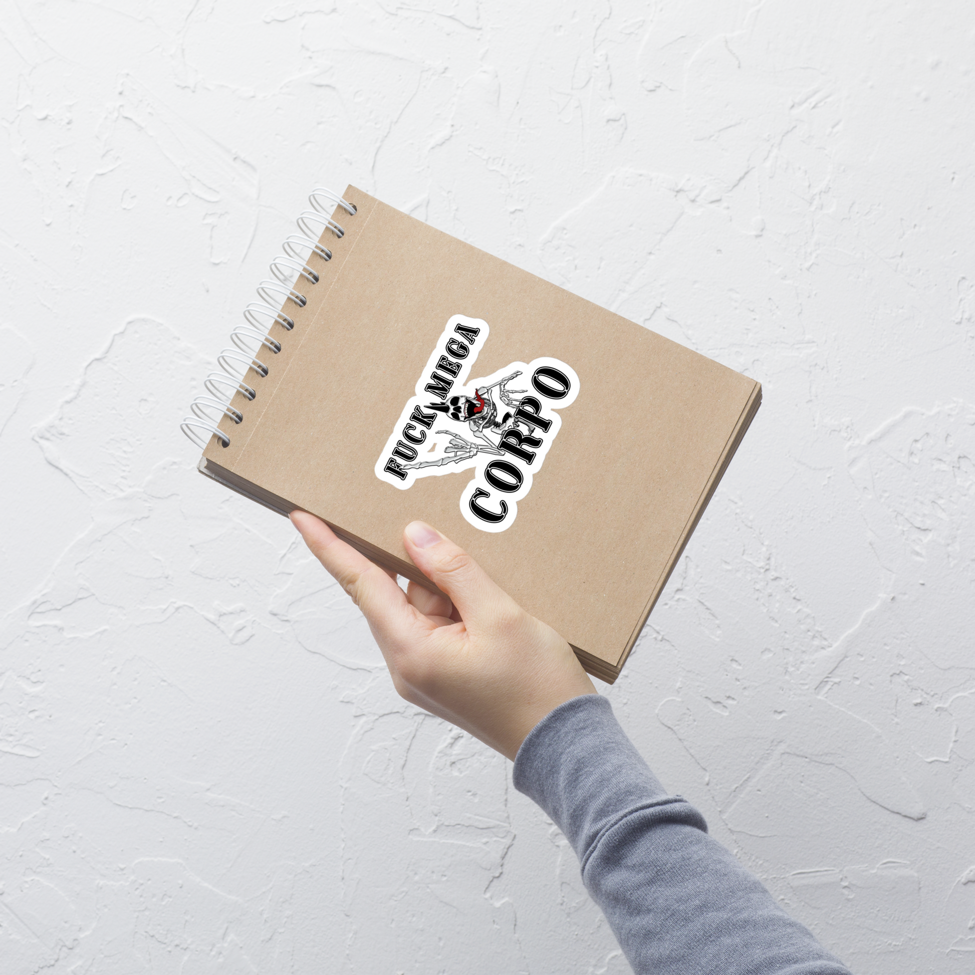 Hand holding a spiral-bound notebook with a sticker with the text 'FUCK MEGA CORPO' and a graphic of a chrome skeleton with a mohawk on a light gray background