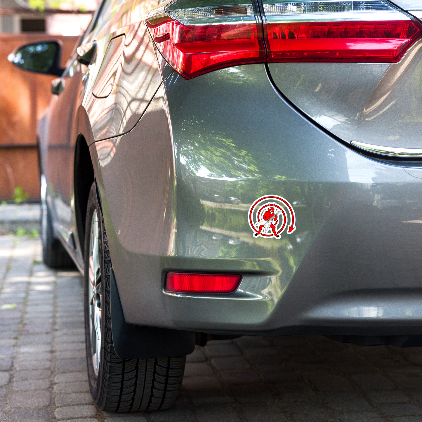 Car's rear end with a sticker depicting a cartoonish red devil playing a guitar, with a predominantly red and white color scheme on a blurred background