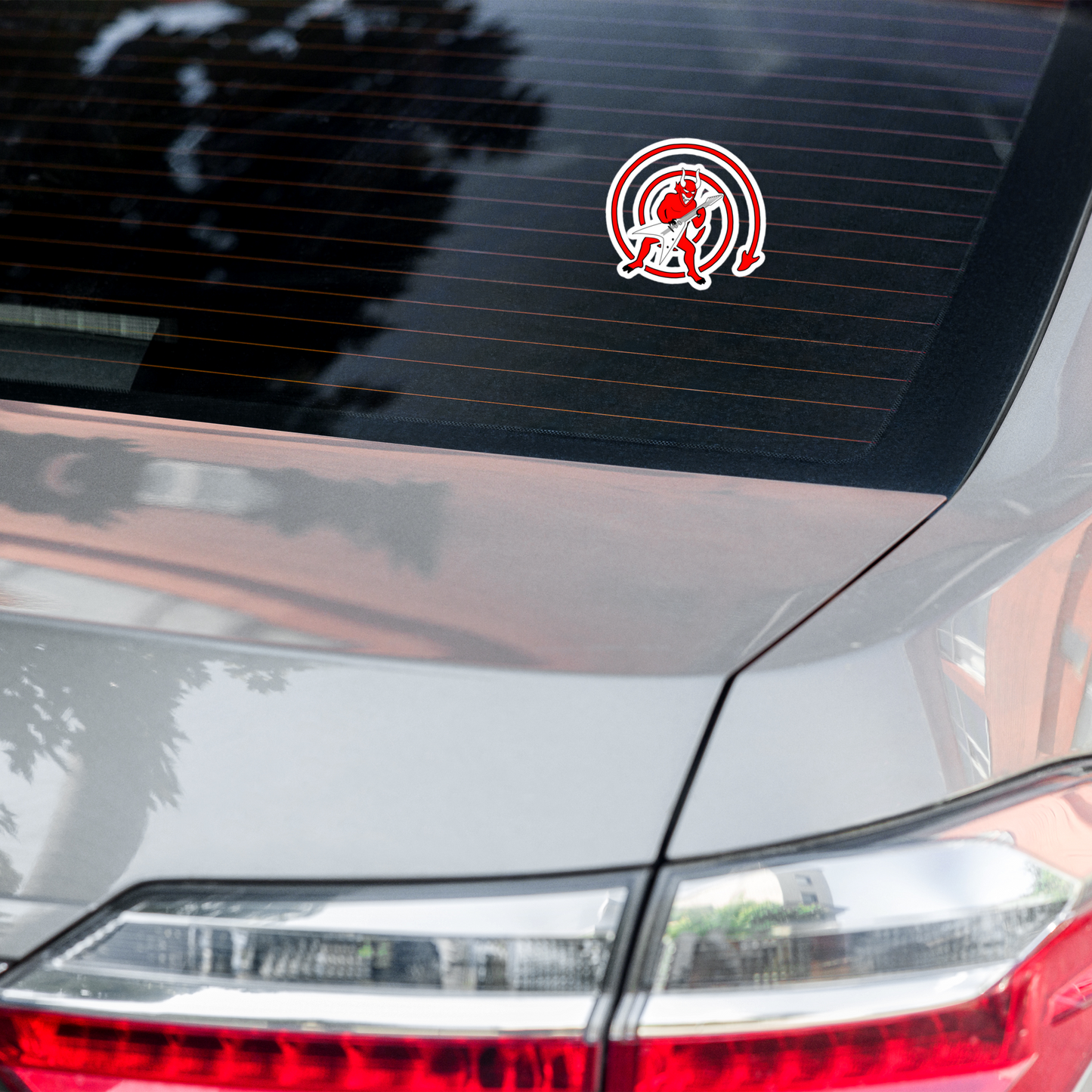 Car sticker depicting a cartoonish red devil playing a guitar, with a predominantly red and white color scheme on a car's rear window.