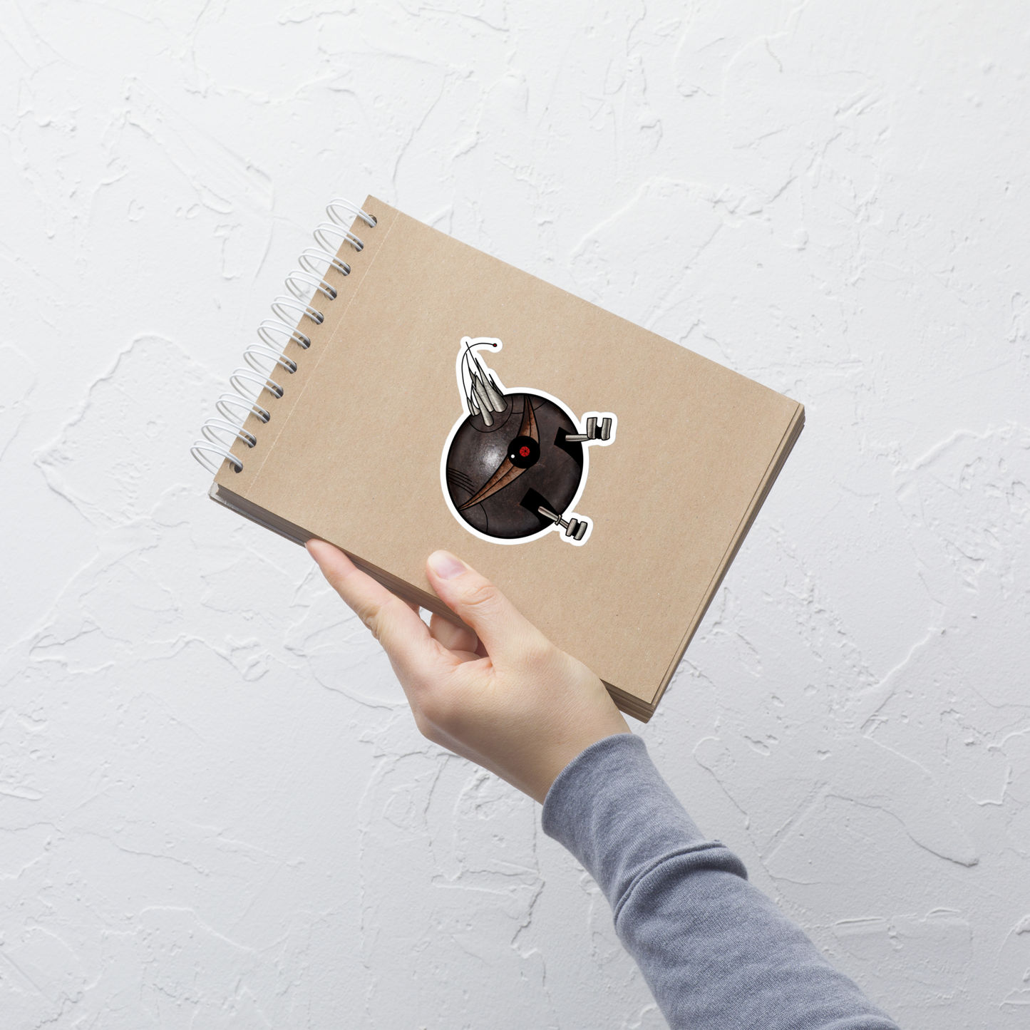 A hand holding a brown kraft paper spiral notebook against a textured white wall. The notebook features a die-cut sticker of a black spherical robot with a red center