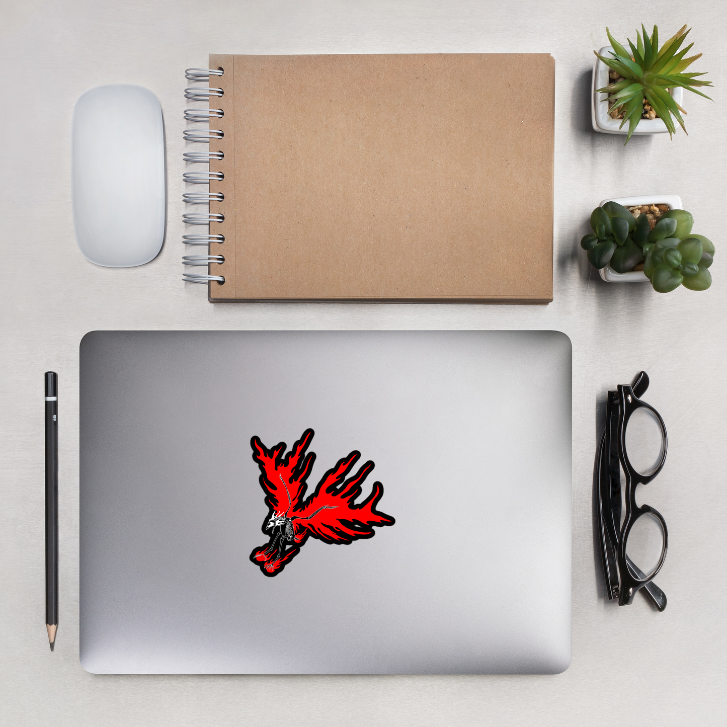 Laptop with a red skeletal eagle design on a desk with a notebook, mouse, pencil, glasses, and plants.
