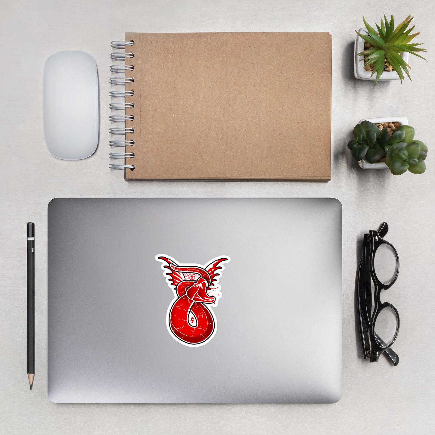 Laptop with a red snake sticker on a desk with a notebook, mouse, pencil, and glasses.