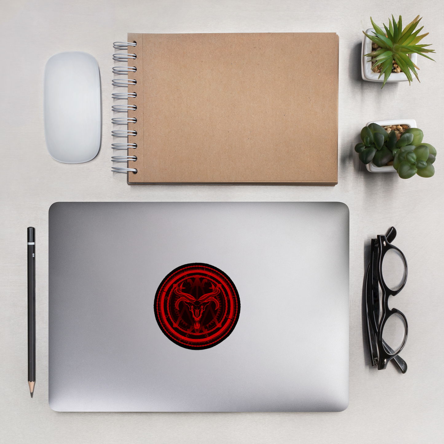 Laptop with a red circular design on a desk with a notebook, mouse, pencil, and glasses.