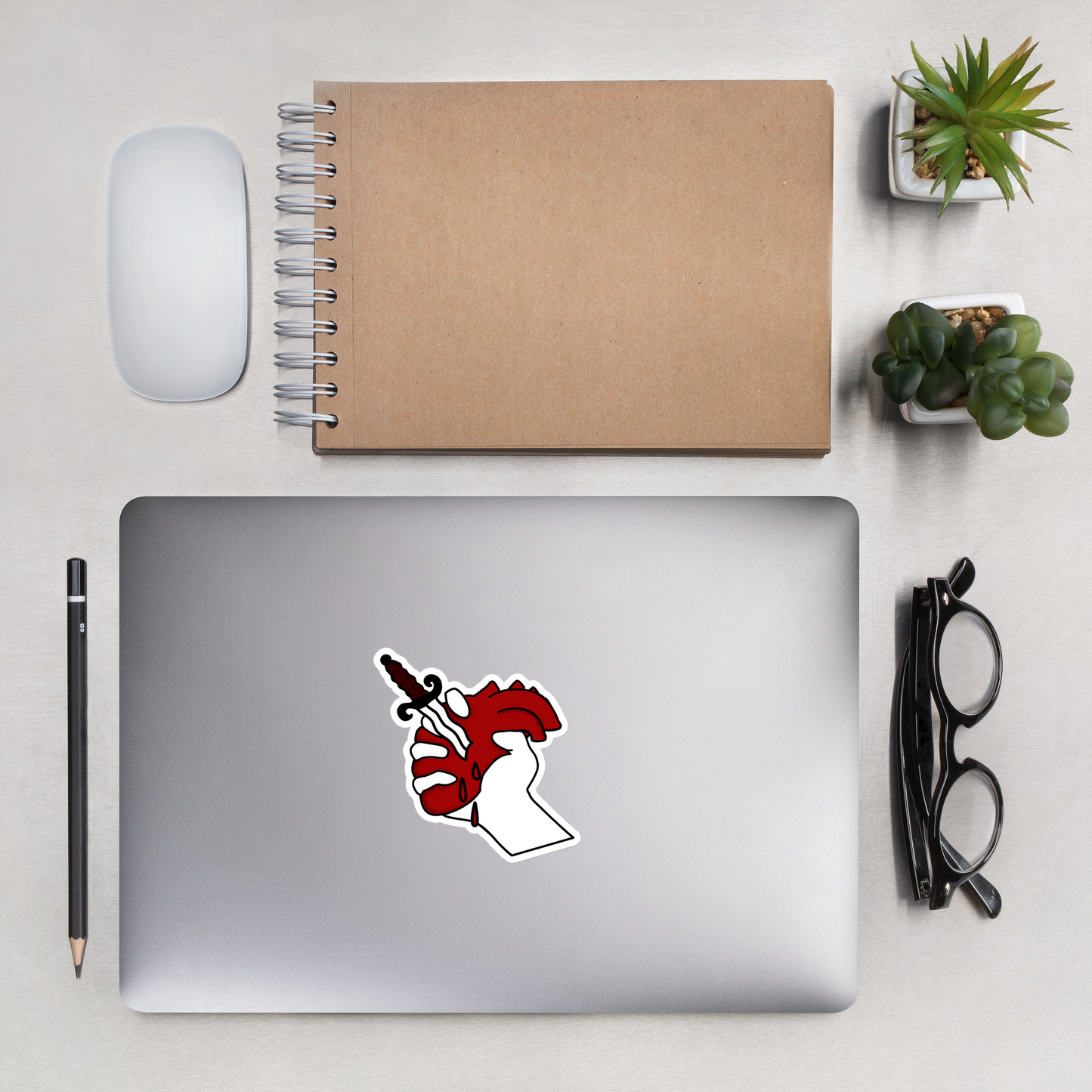 Laptop with a heart and dagger sticker on a desk with a notebook, mouse, pencil, and glasses.