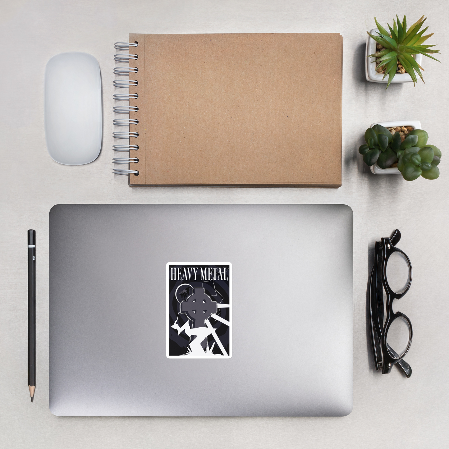 Laptop with a 'Heavy Metal' sticker on a desk with notebook, mouse, pencil, and plants.