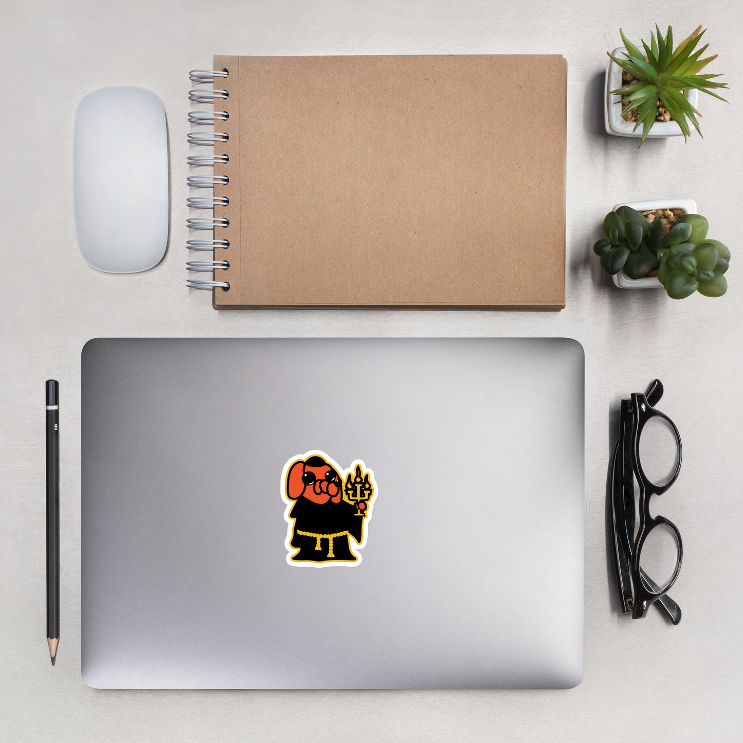 Laptop with a sticker of an orange cartoon dog, black robe, holding a candelabra on a white background on a desk with a notebook, mouse, pencil, and glasses.