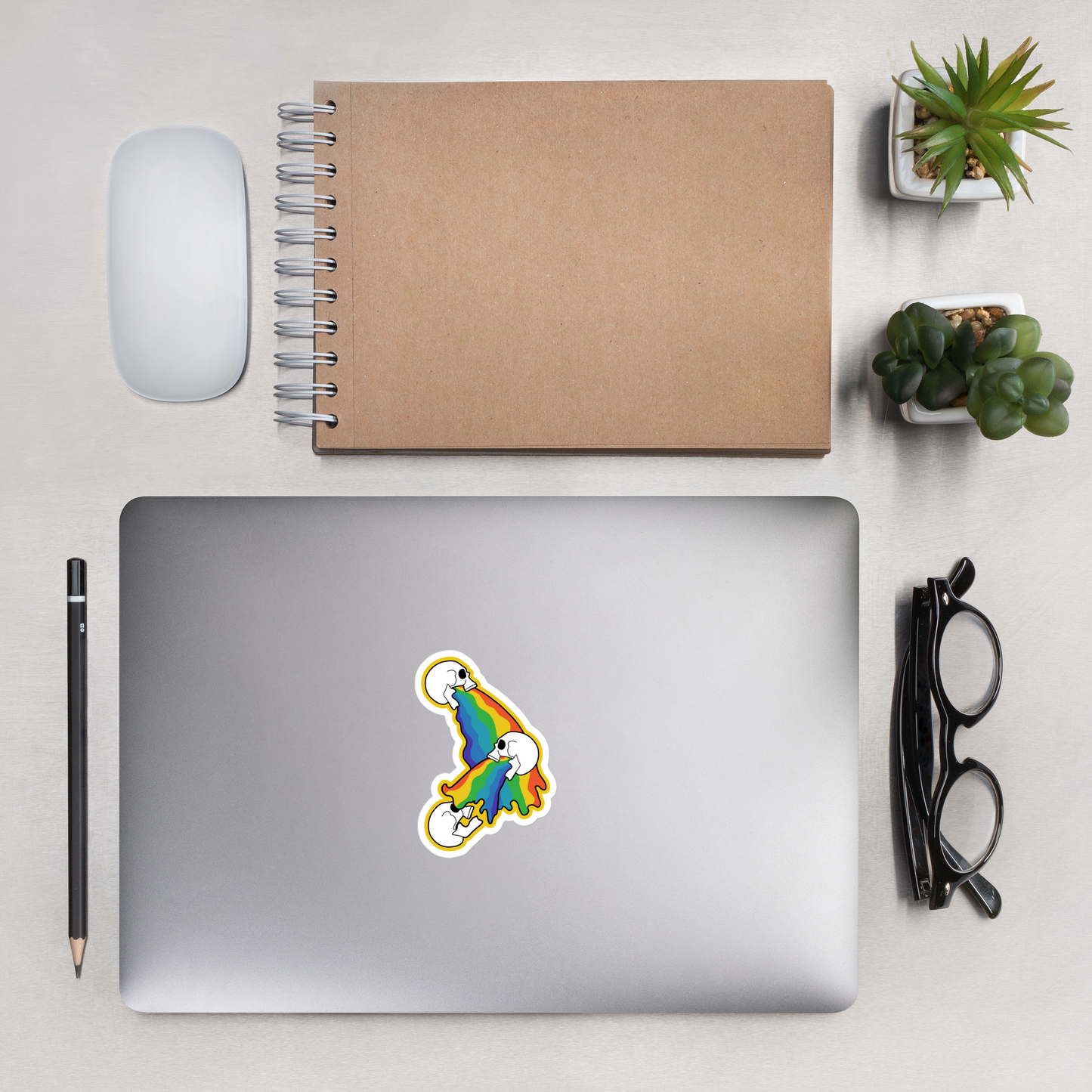 Laptop with a colorful rainbow skull sticker on a desk with a notebook, mouse, pencil, and glasses.