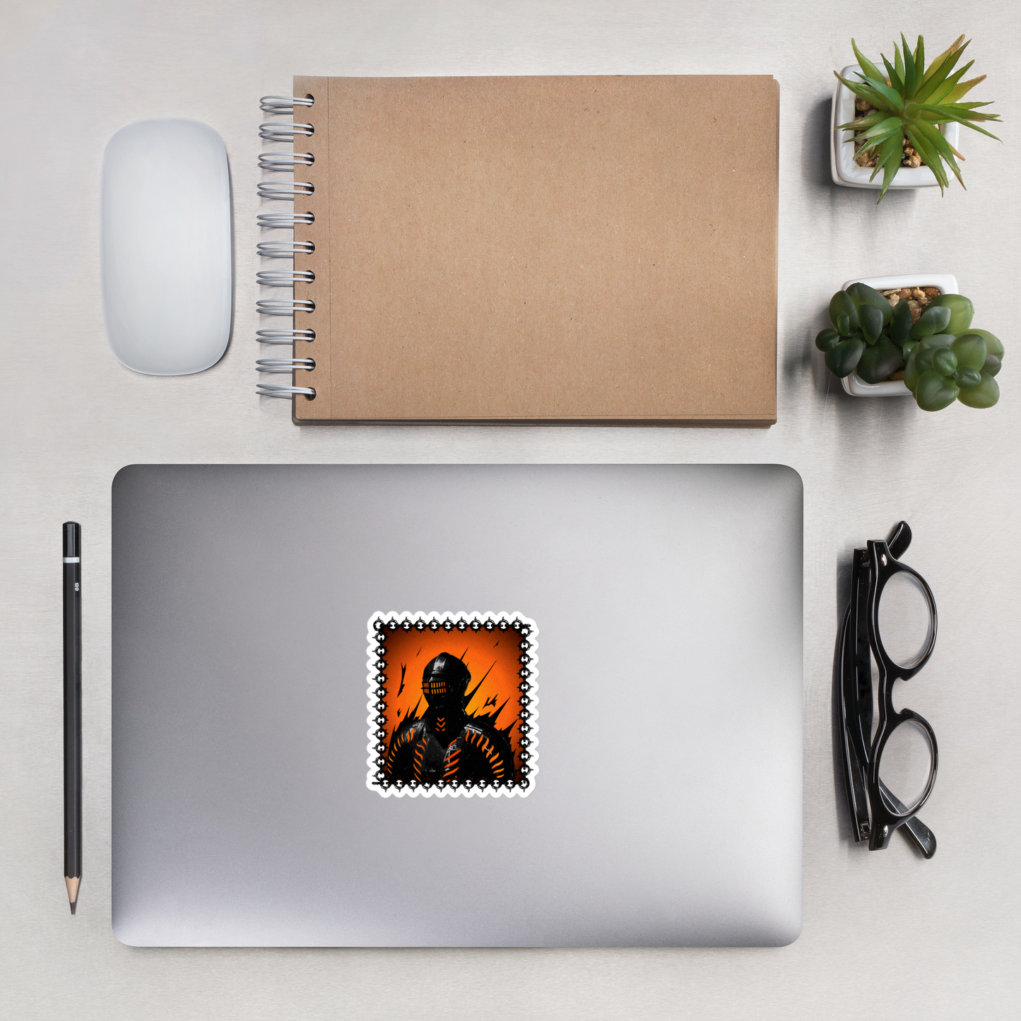 Laptop with a stylized sticker of a knight in a helmet and armor with orange accents on a decorative border. on a desk with a notebook, mouse, pencil, and plants.