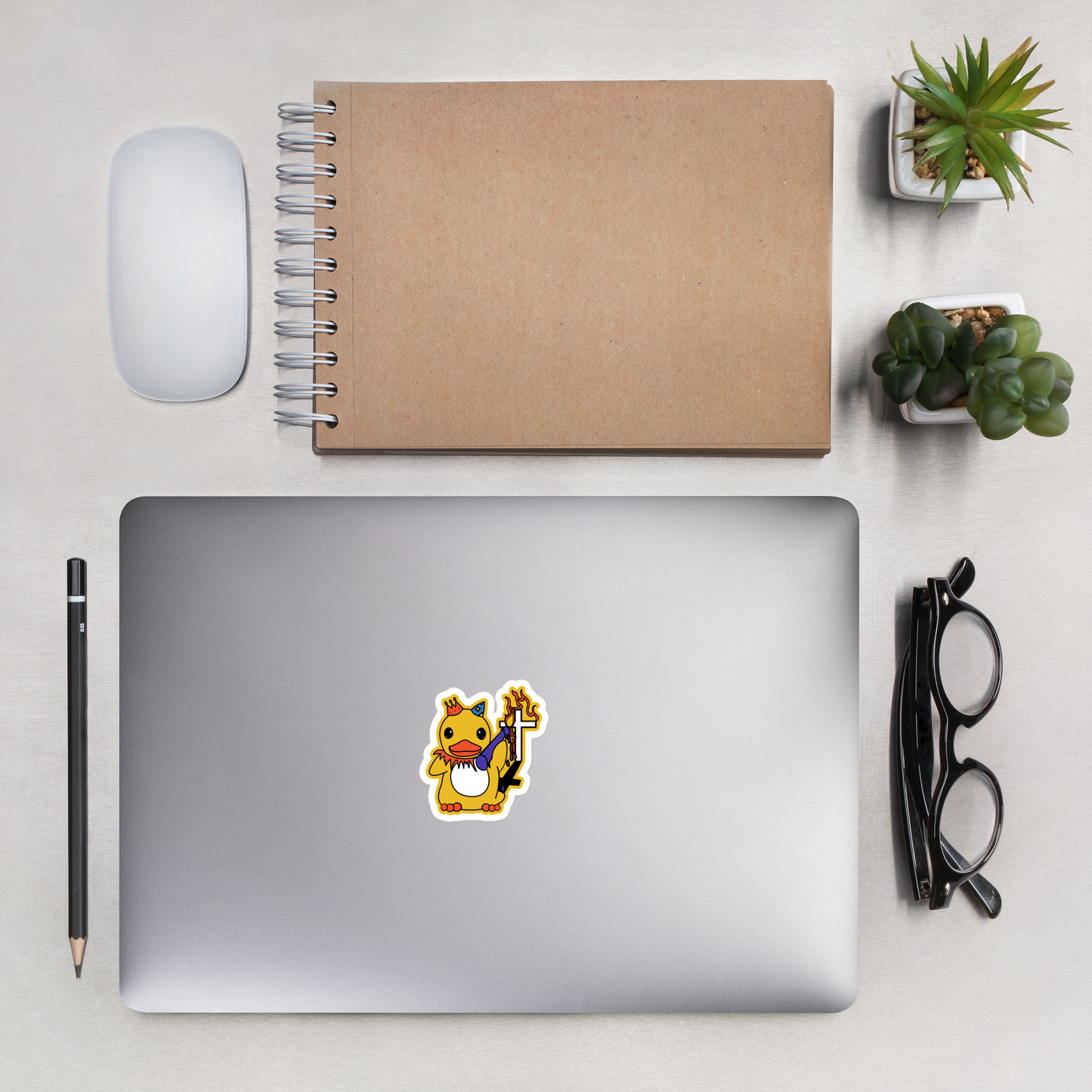 Laptop with a sticker of a colorful duck holding a flaming candle in front of a cross grave on a desk with a notebook, mouse, pencil, and plants.