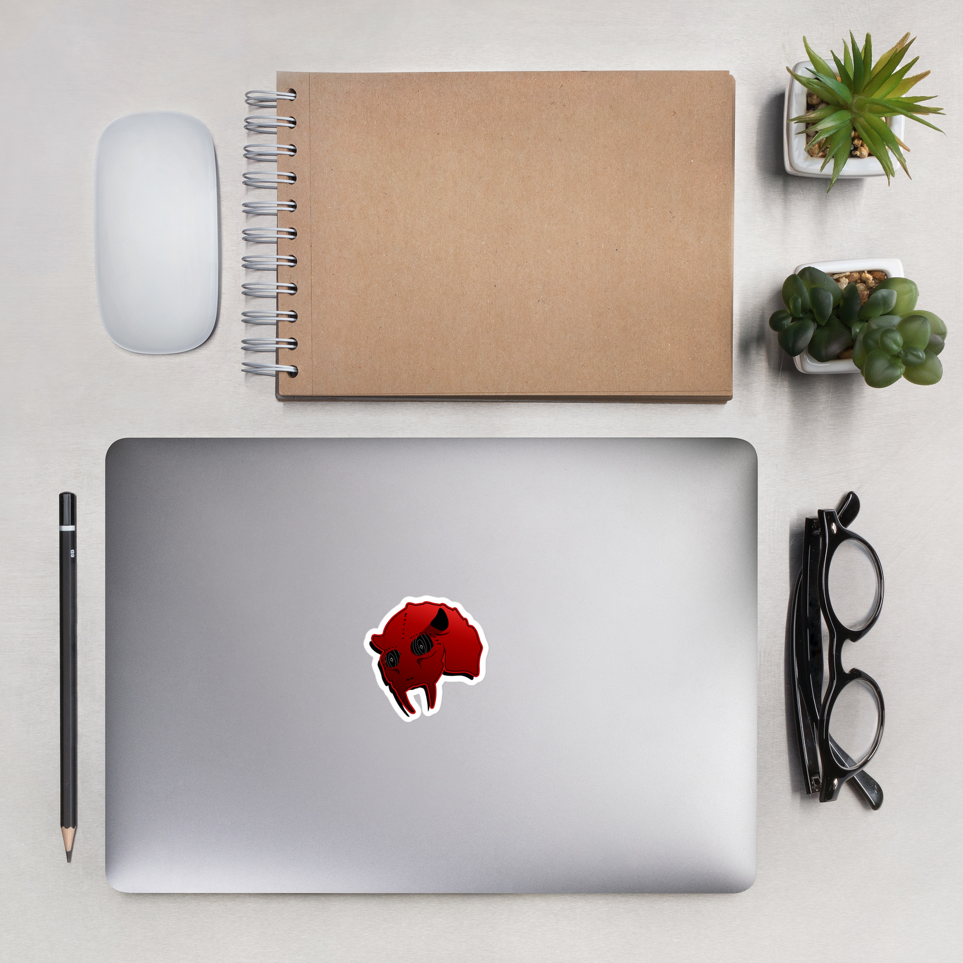 Laptop with a red heavy emtal sticker on a desk with a notebook, mouse, pencil, and glasses.