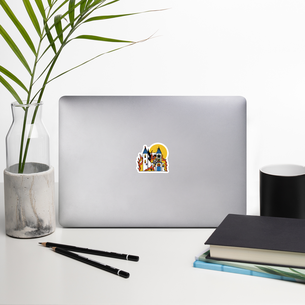 Laptop with a sticker of a silly sticker with a colorful illustration of a giant monster attacking a flaming church full of teddy bears on a desk with a plant, pencils, and books.