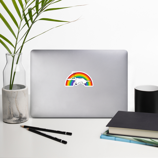 Laptop with a die-cut sticker featuring a white cartoon creature with a long tail, set against a large rainbow arch on a plain white background on a desk with a plant, pencils, and notebook.