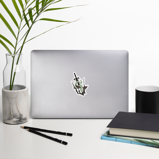 Laptop with a die-cut vinyl sticker featuring a stylized green snake wrapped around a black sword, shown on a plain white background on a desk with a plant, mug, and books.