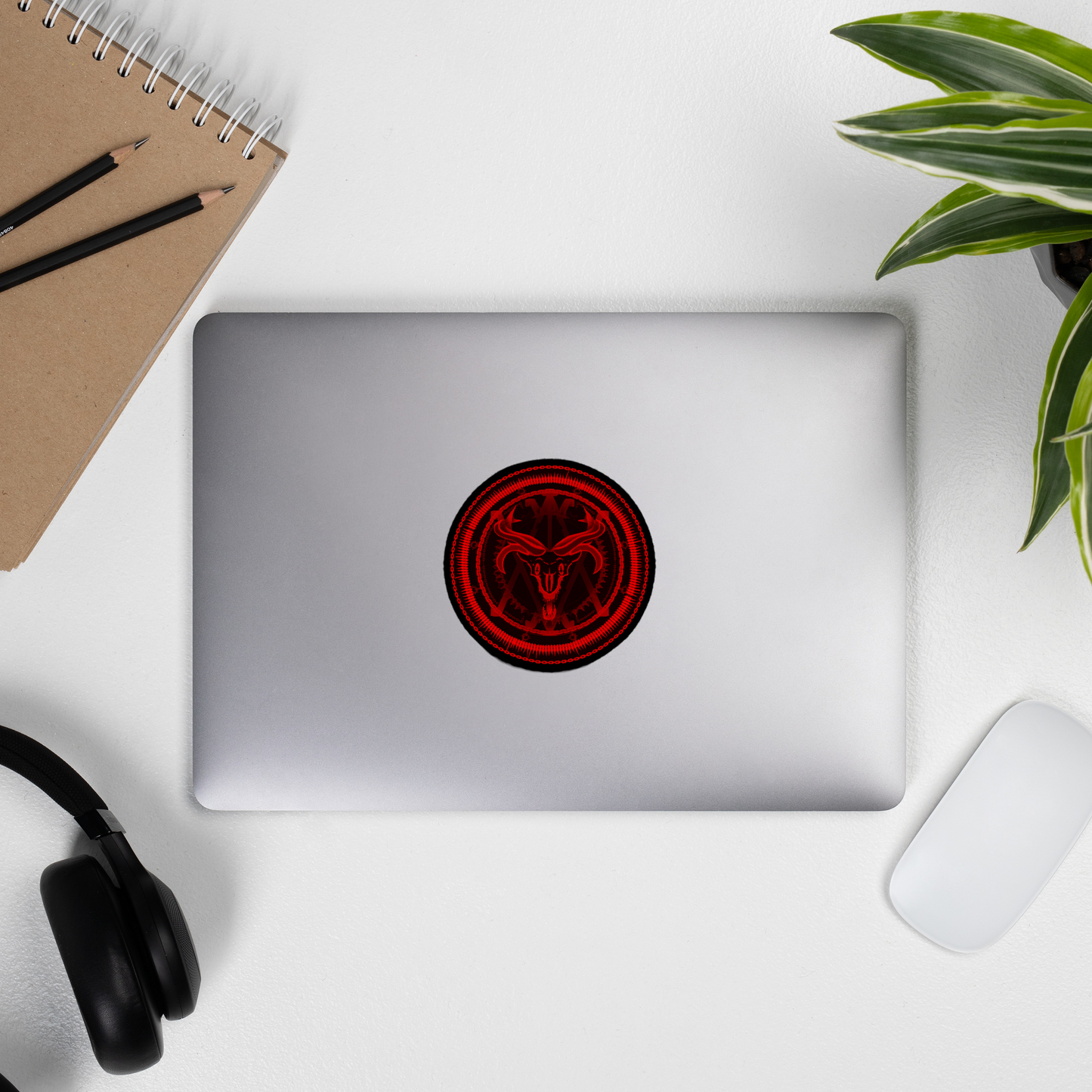 Laptop with a red and black circular demonic design on a white surface with a plant and office supplies.
