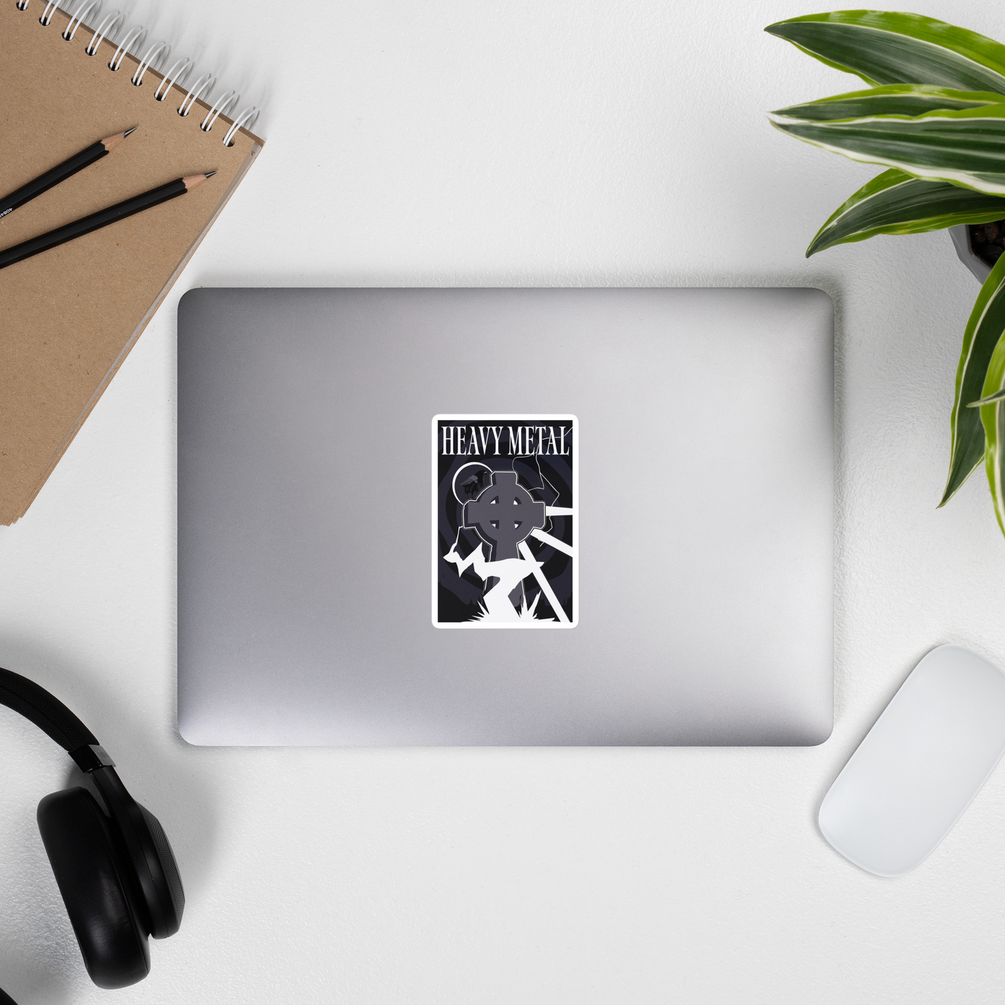 Laptop with a 'Heavy Metal' sticker on a desk with a notebook, pencils, headphones, and a plant.