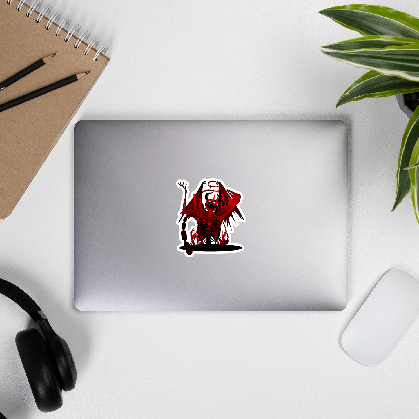 Laptop with a red and black skeleton demon sticker on a white surface with a notebook, pencils, and plant.