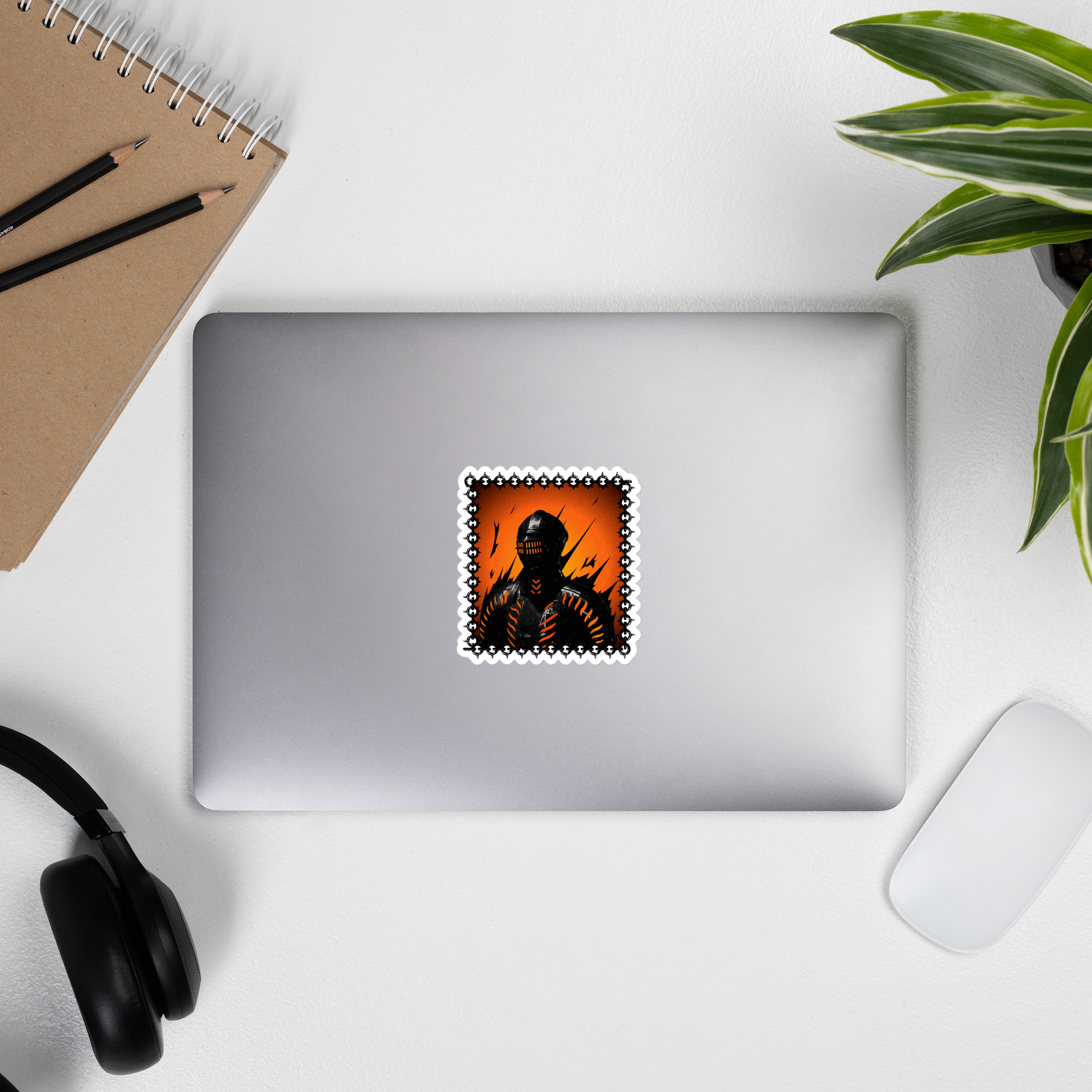 Laptop with a sticker of a stylized sticker of a knight in a helmet and armor with orange accents on a desk with a notebook, pencils, headphones, and a plant.