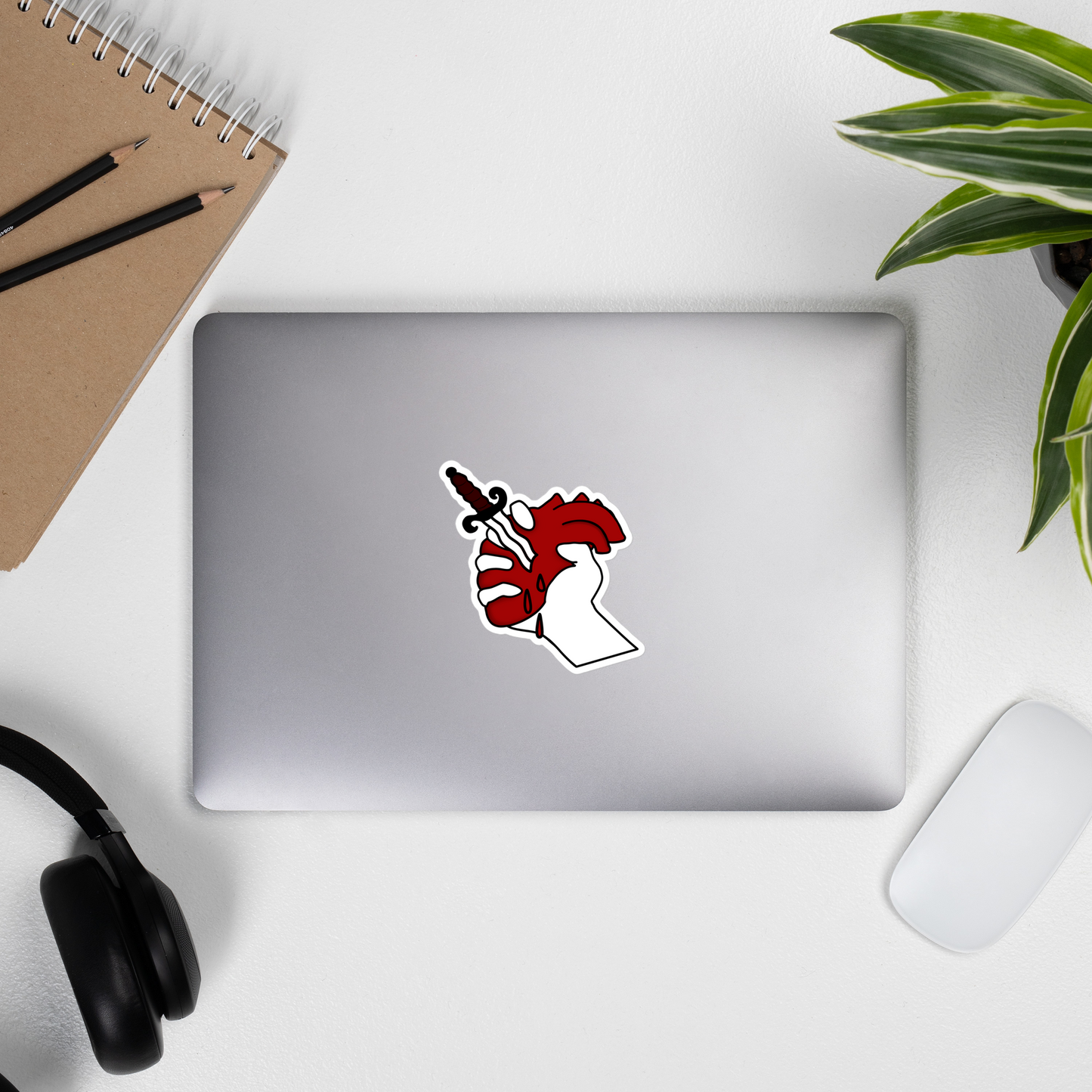 Laptop with a sticker of a heart being held up with a dagger in it on a desk with a notebook, pencils, headphones, and a plant.