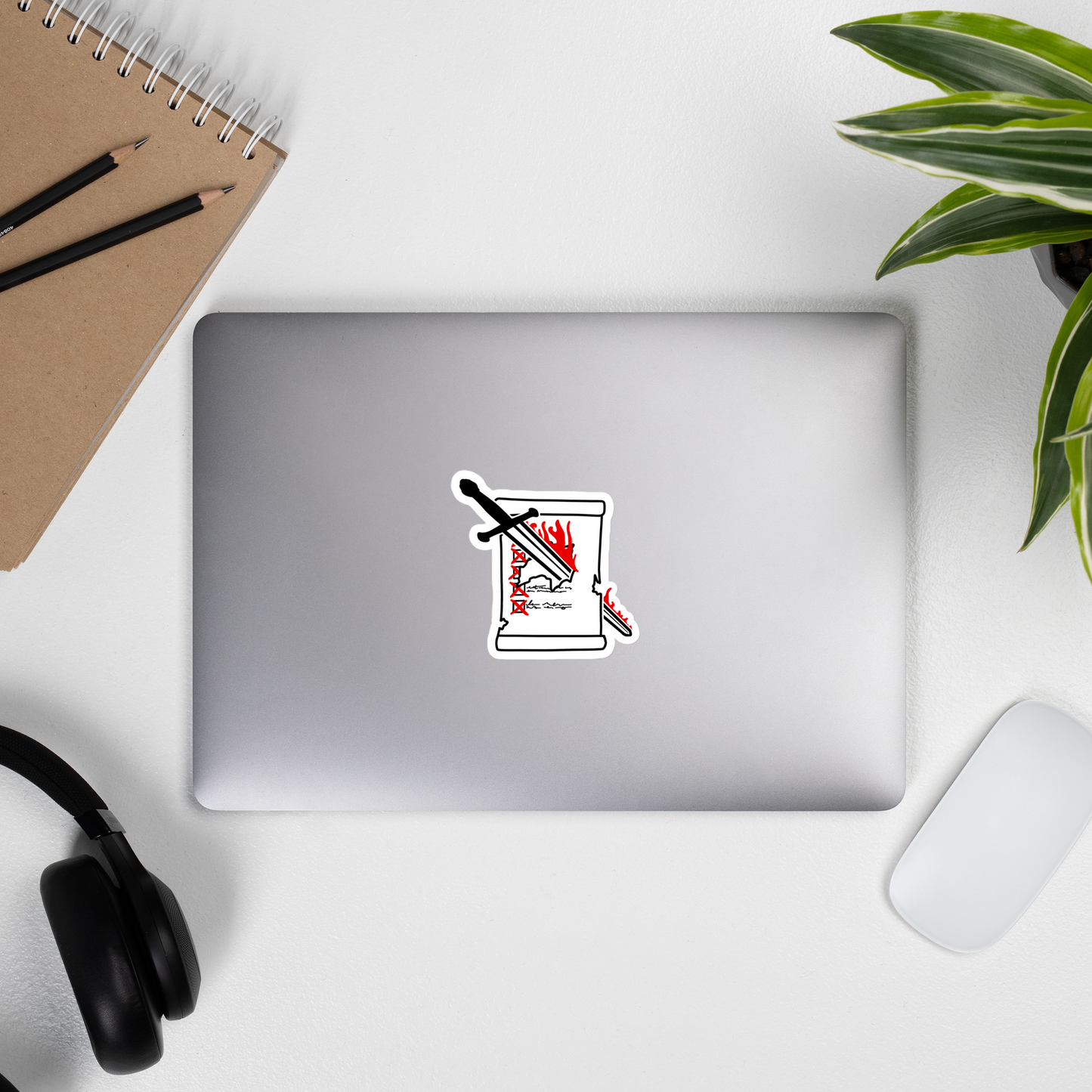 Laptop with a sticker of flaming sword cutting through a checklist scroll on a desk with a notebook, pencils, headphones, and a plant.