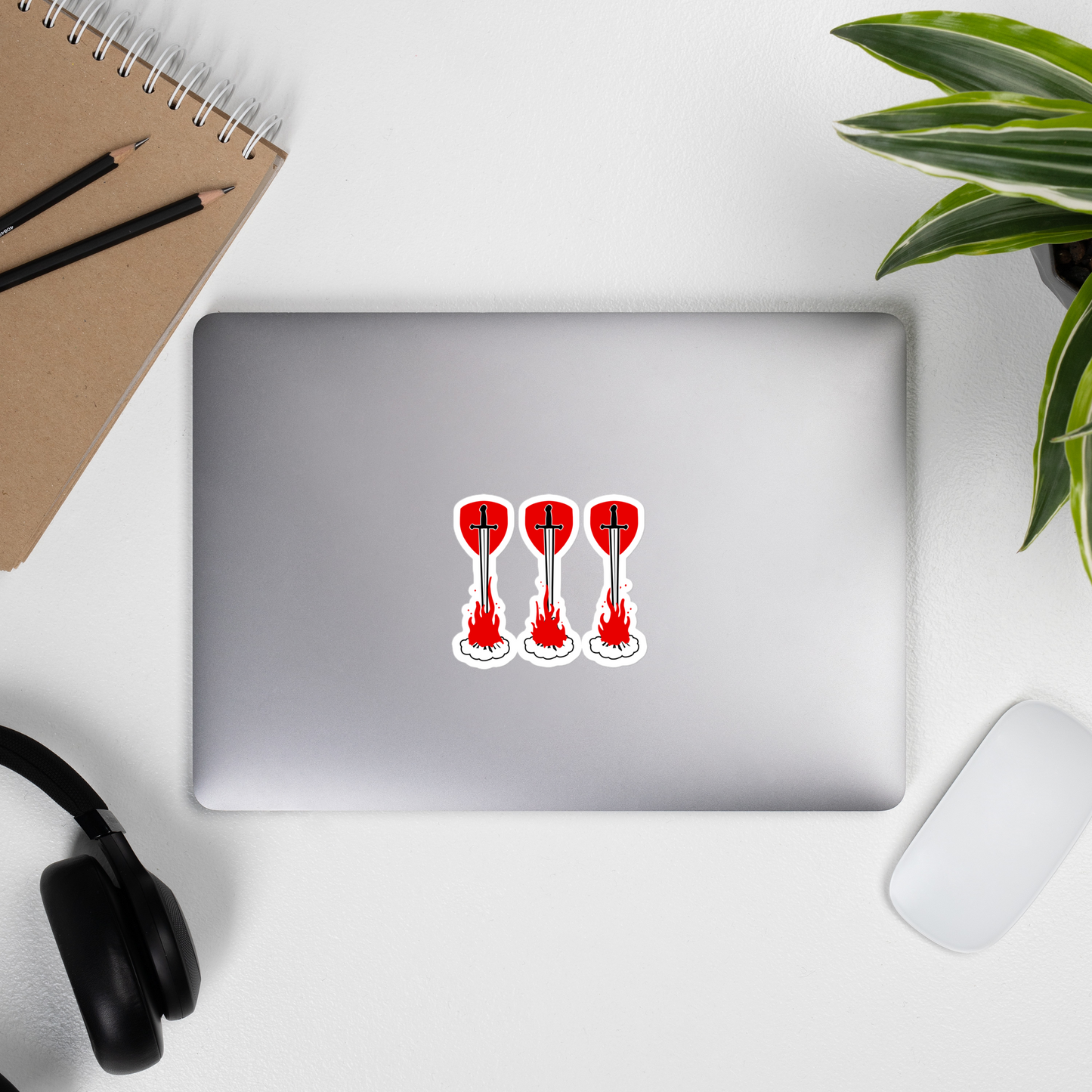 Laptop with sticker of three swords over campfires in front of shields on a desk with a notebook, pencils, and headphones.