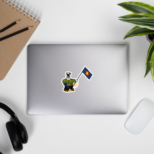 A die-cut sticker of a cartoon knight in green armor holding a blue flag with a yellow star, shown on the back of a silver laptop. The laptop is styled in a flat-lay desk setup with a notebook, headphones, a mouse, and a plant.