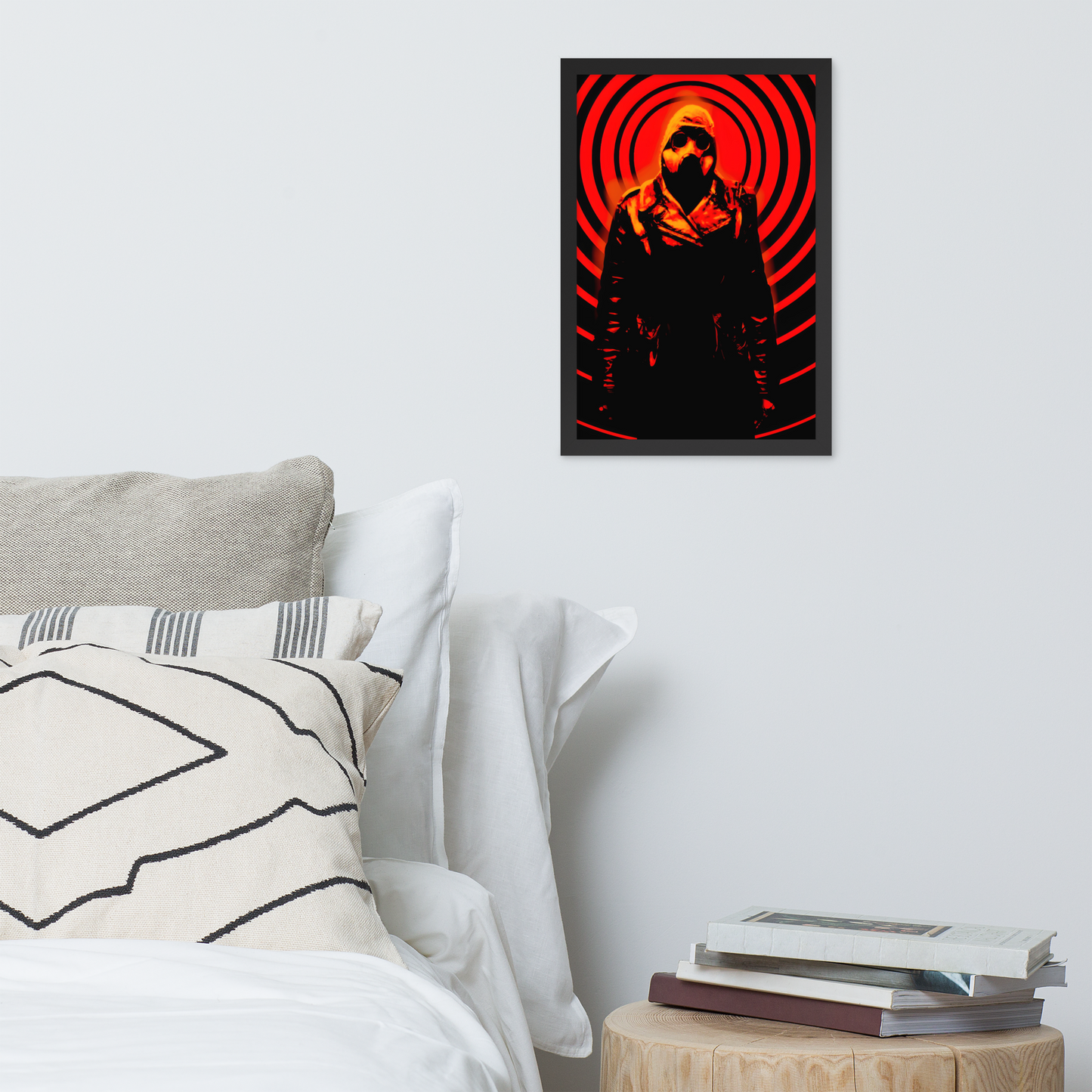 Framed artwork of a mysterious figure in a dark outfit with a hood and a gas mask against an orange and black circular background above a bed with pillows and books on a nightstand.