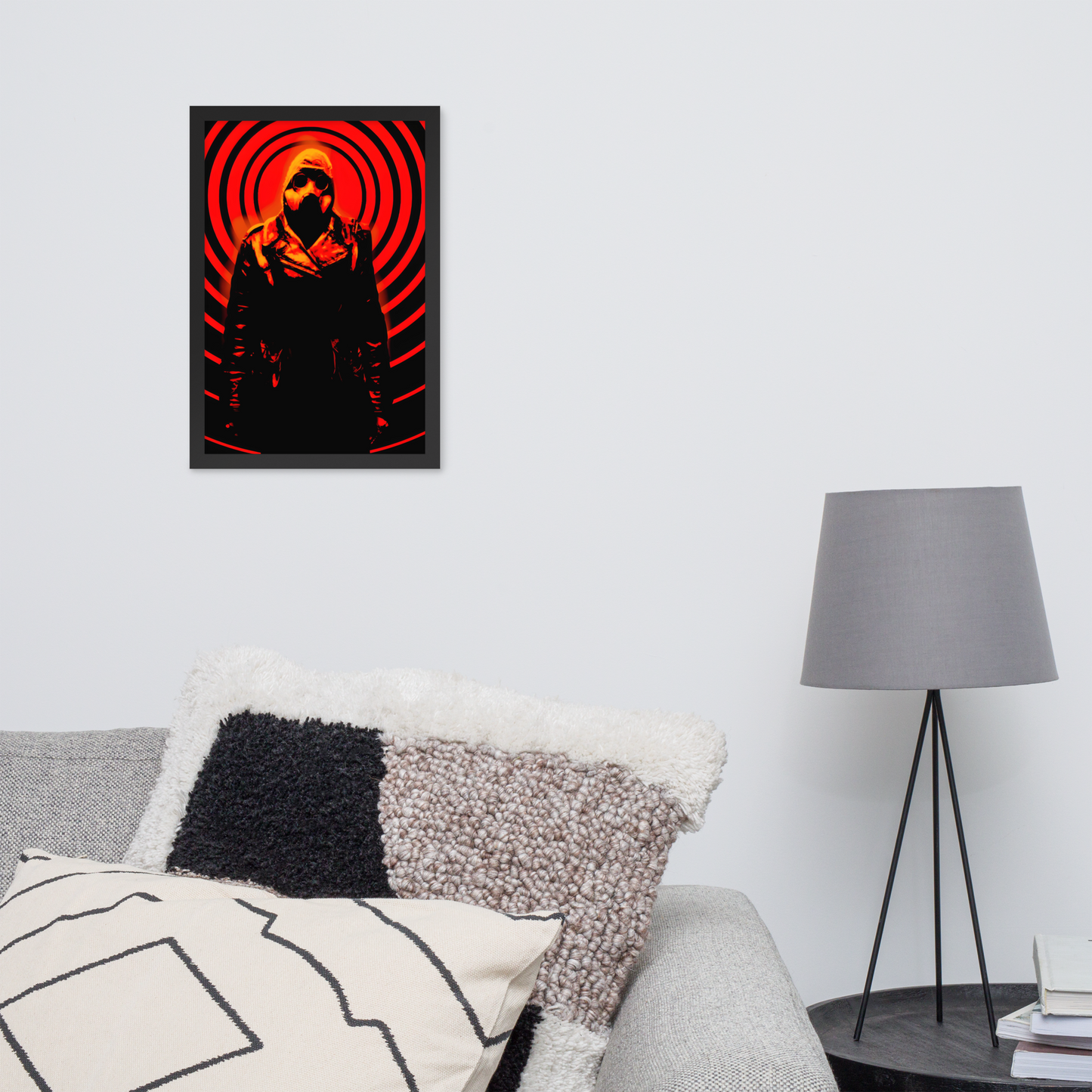 12x18 framed artwork of a mysterious figure in a dark outfit with a hood and a gas mask against an orange and black circular background on a wall above a sofa with decorative pillows and a lamp.