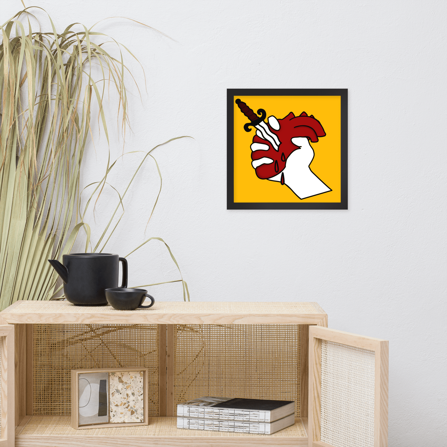 Framed artwork of a hand holding up a heart with a dagger running through it on a yellow background above a wooden shelf with decor items.
