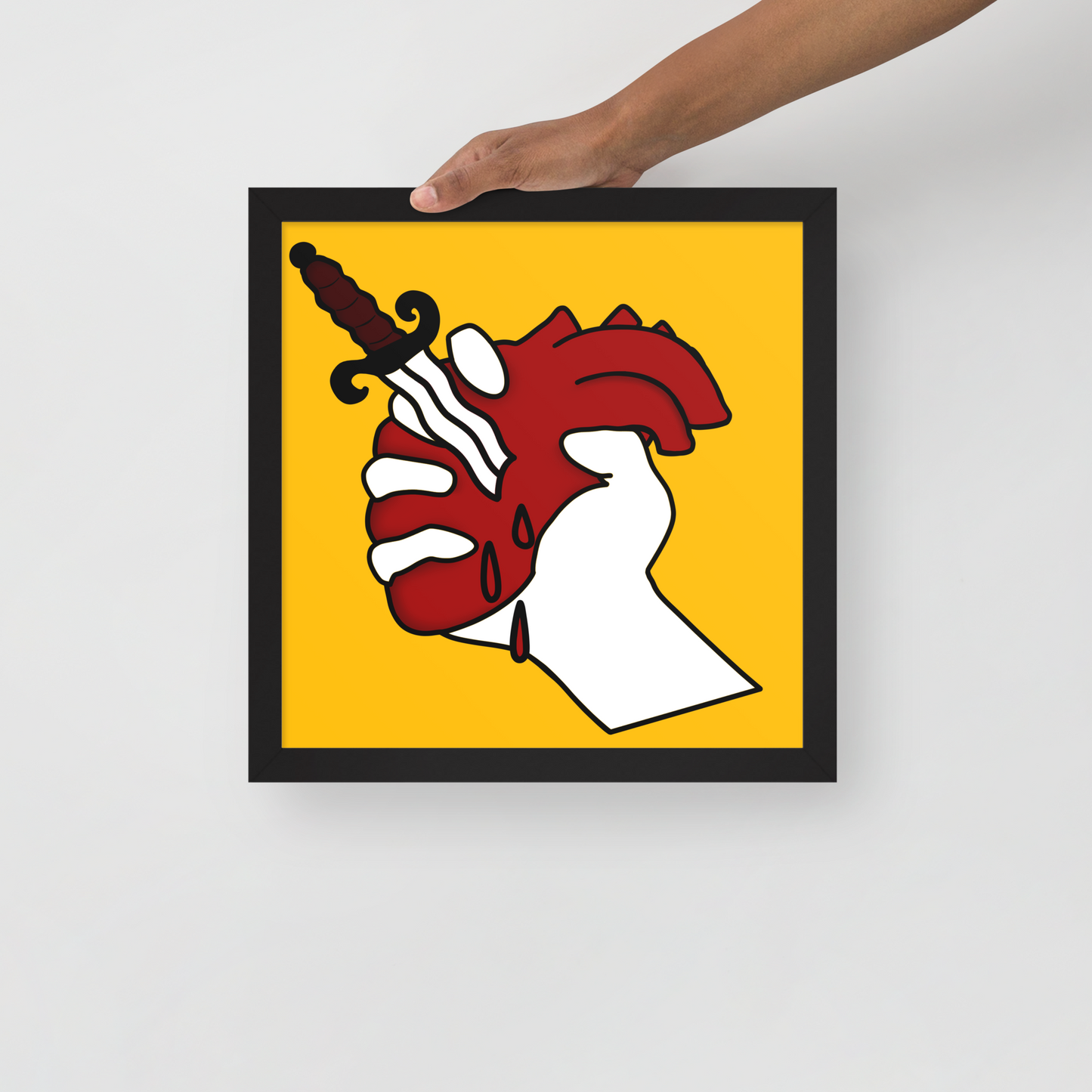 Framed artwork of a hand holding a dagger with a red heart on a yellow background, held by a hand against a white background.