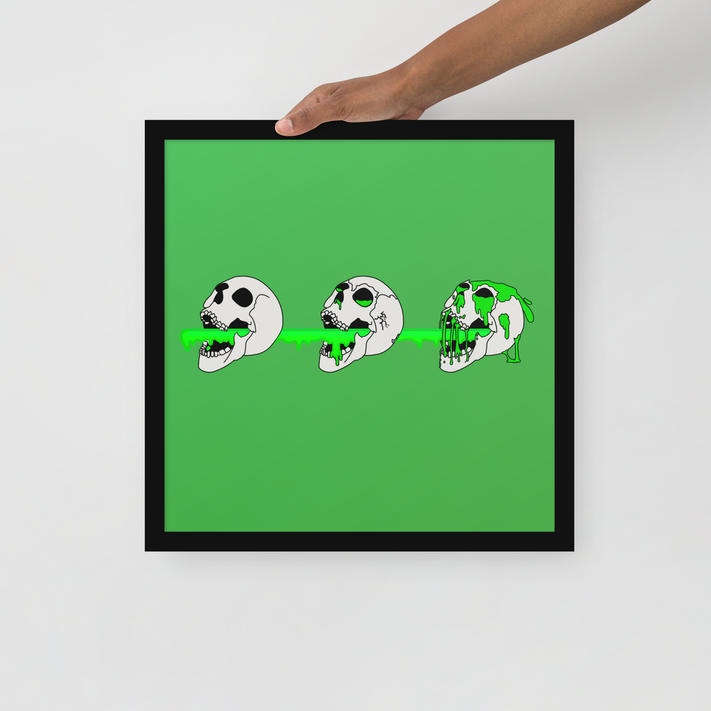 Framed artwork with a green background and black frame, featuring three cartoonish skull designs.