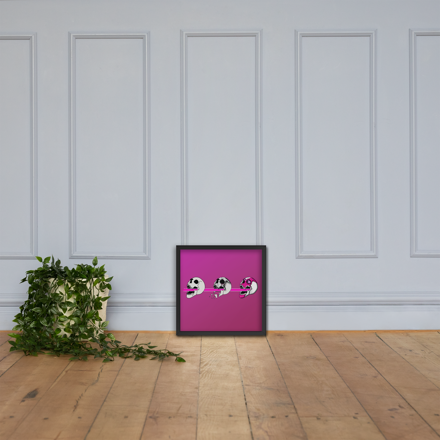 Framed artwork with a pink background and black frame, featuring three stylized cyberpunk skull designs shooting a laser with purple and black details against a white paneled wall by a plant.